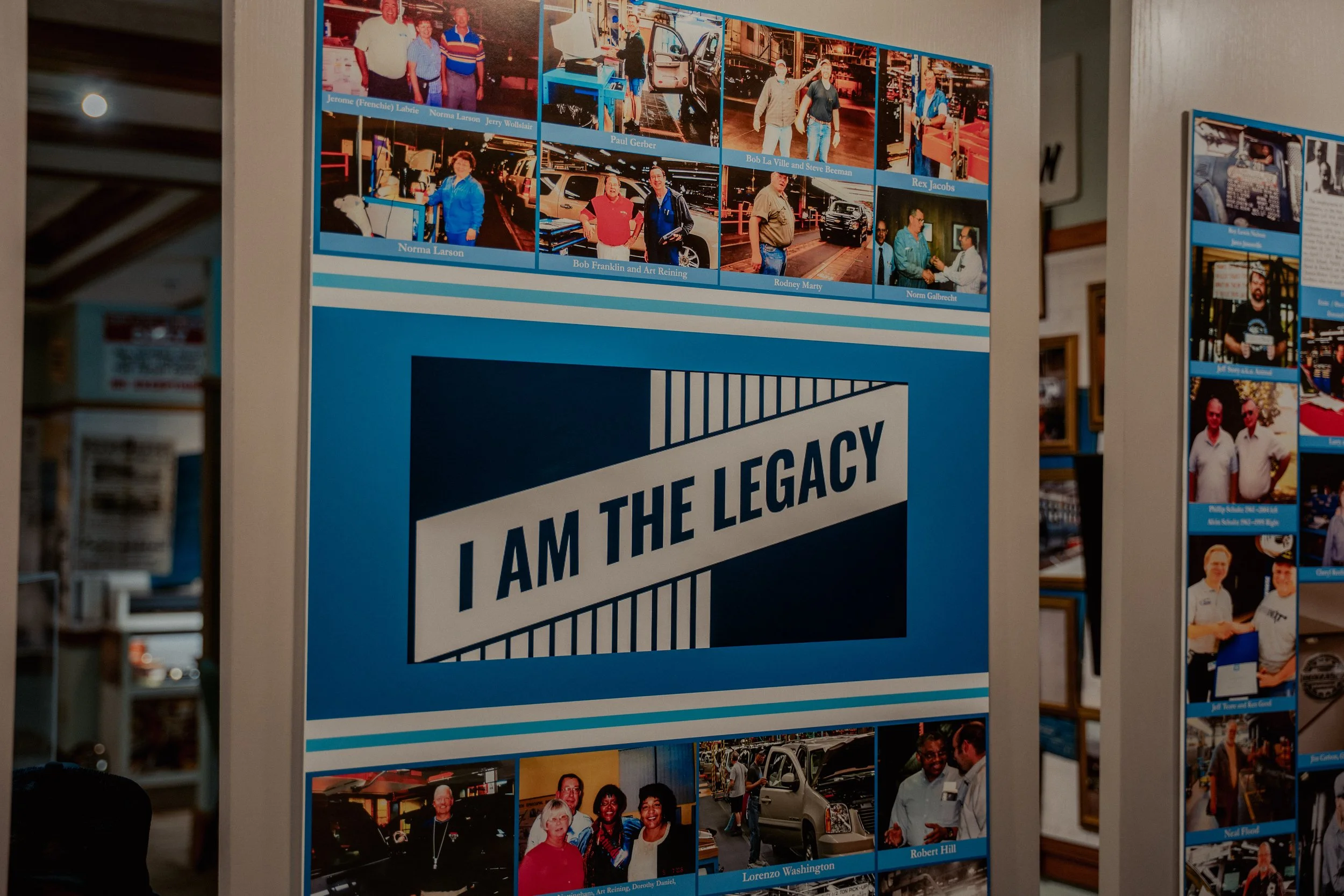 A display board featuring photographs of various people, each with a name label, and a large sign that reads "I AM THE LEGACY." The photos depict events and individuals associated with a community or organization.