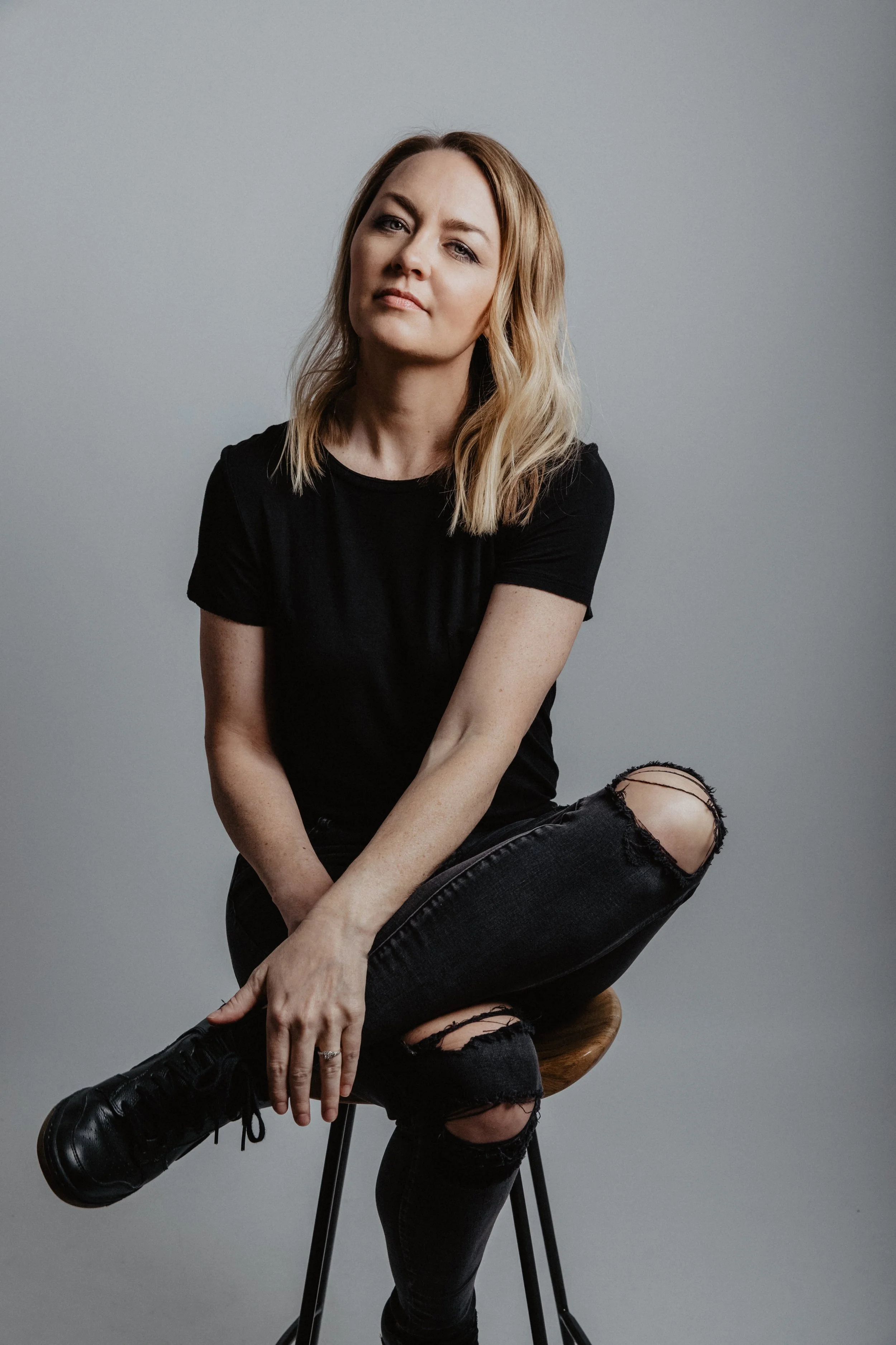 Portrait of a woman with blonde hair, wearing a black t-shirt and ripped black jeans, sitting on a stool with a neutral gray background.