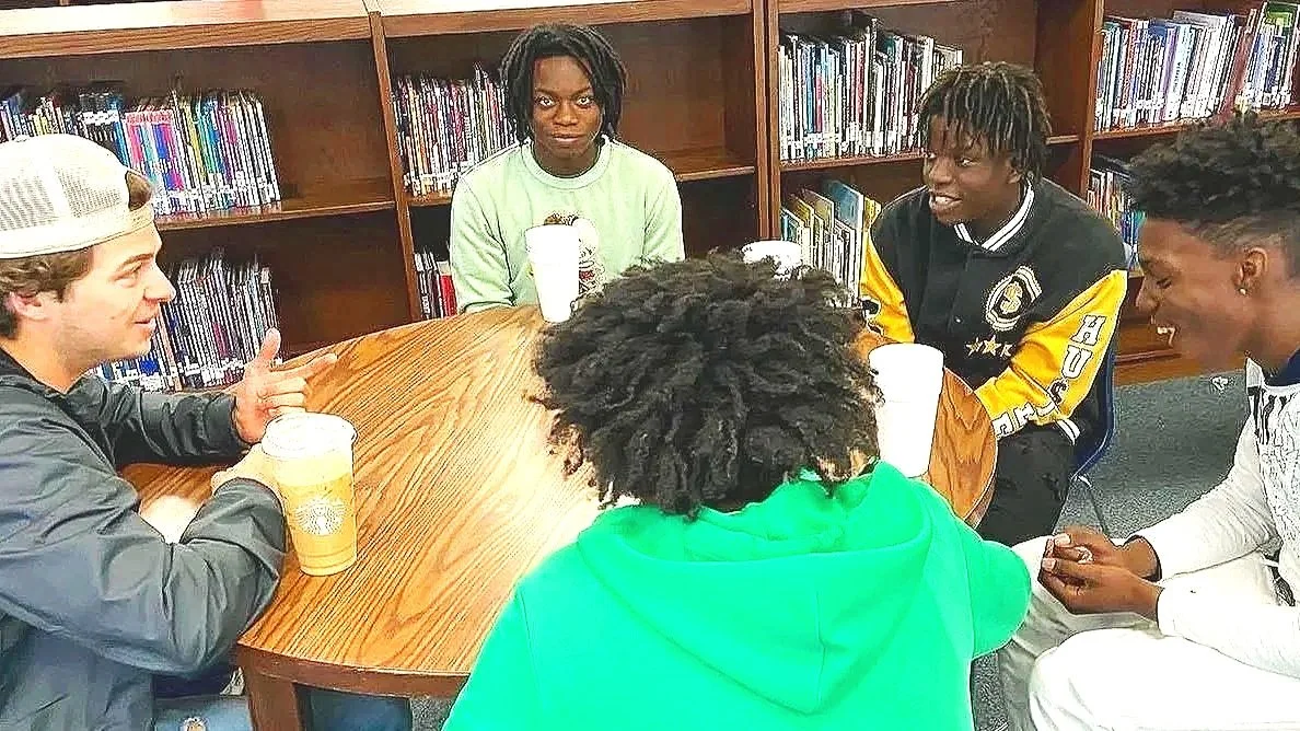 A group of five young people sitting around a table in a library, having a conversation and enjoying beverages.