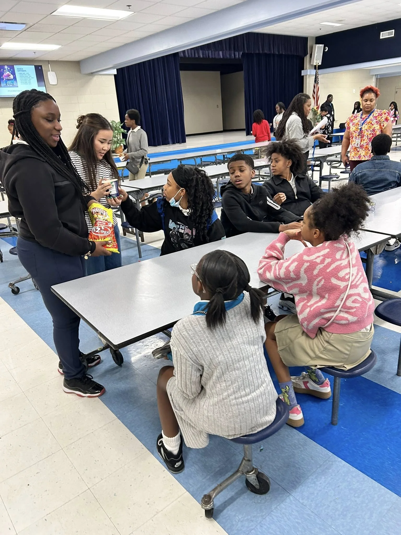 Group of children and adults in a school cafeteria or multipurpose room, some sitting at tables and others standing, engaged in conversations and activities.