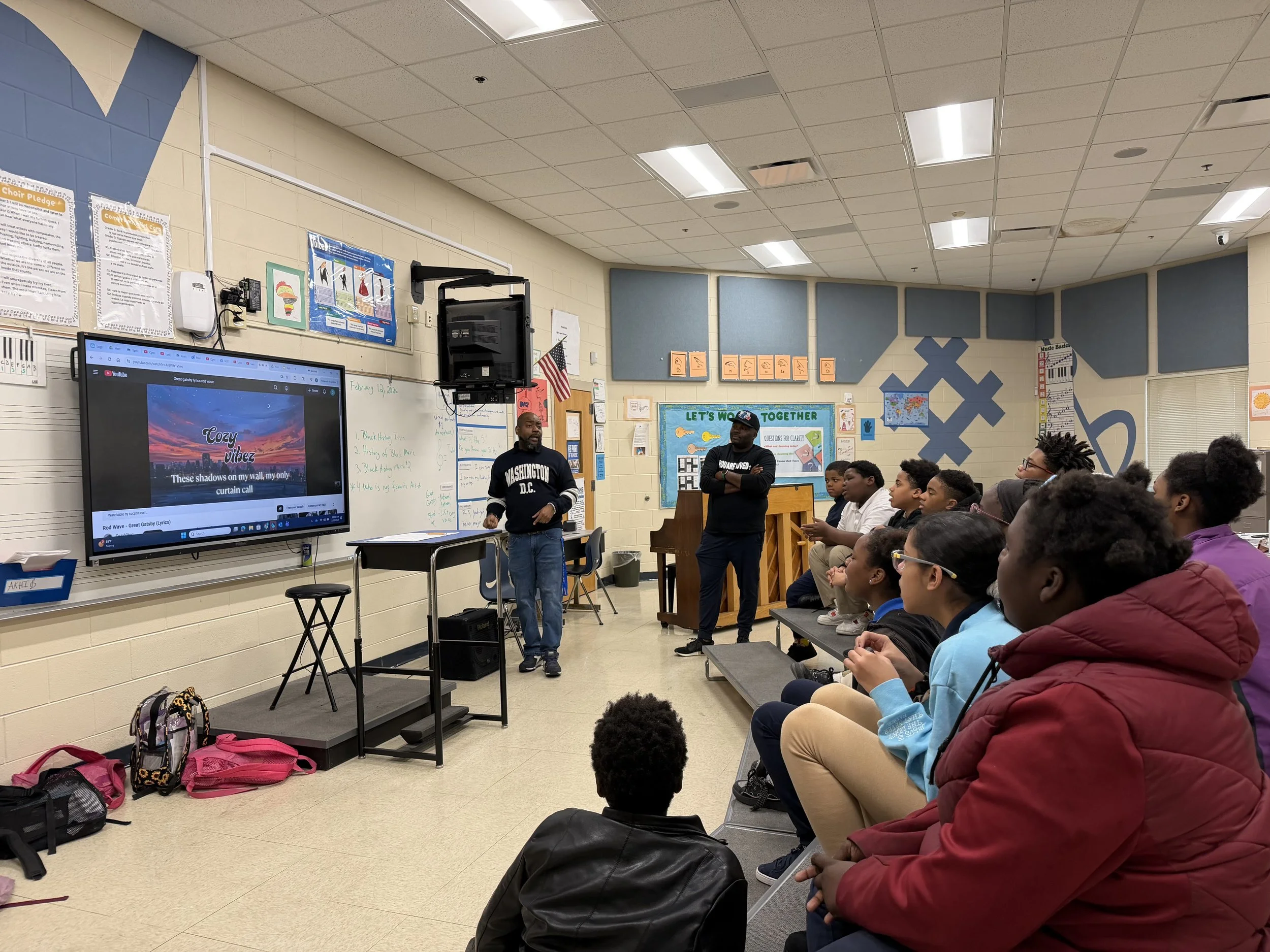 A classroom with students seated on a bench, watching a presentation on a large screen. Two men stand at the front, one near a whiteboard and the other next to a piano. The classroom has colorful decorations and posters on the walls.