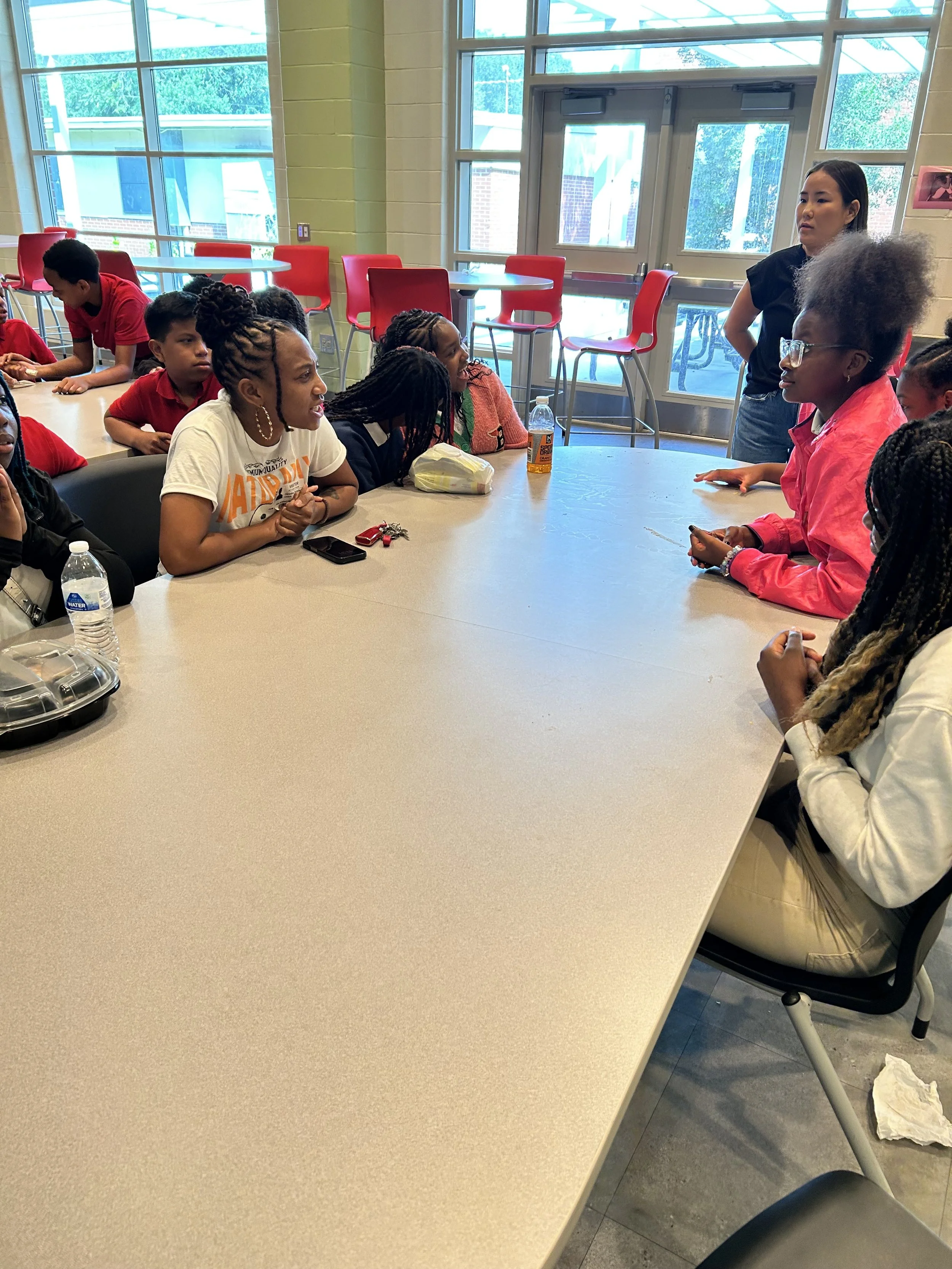 A group of children sitting around a large table in a classroom, engaged in a discussion with a woman standing at the end of the table. The room has large windows, red chairs, and natural daylight.