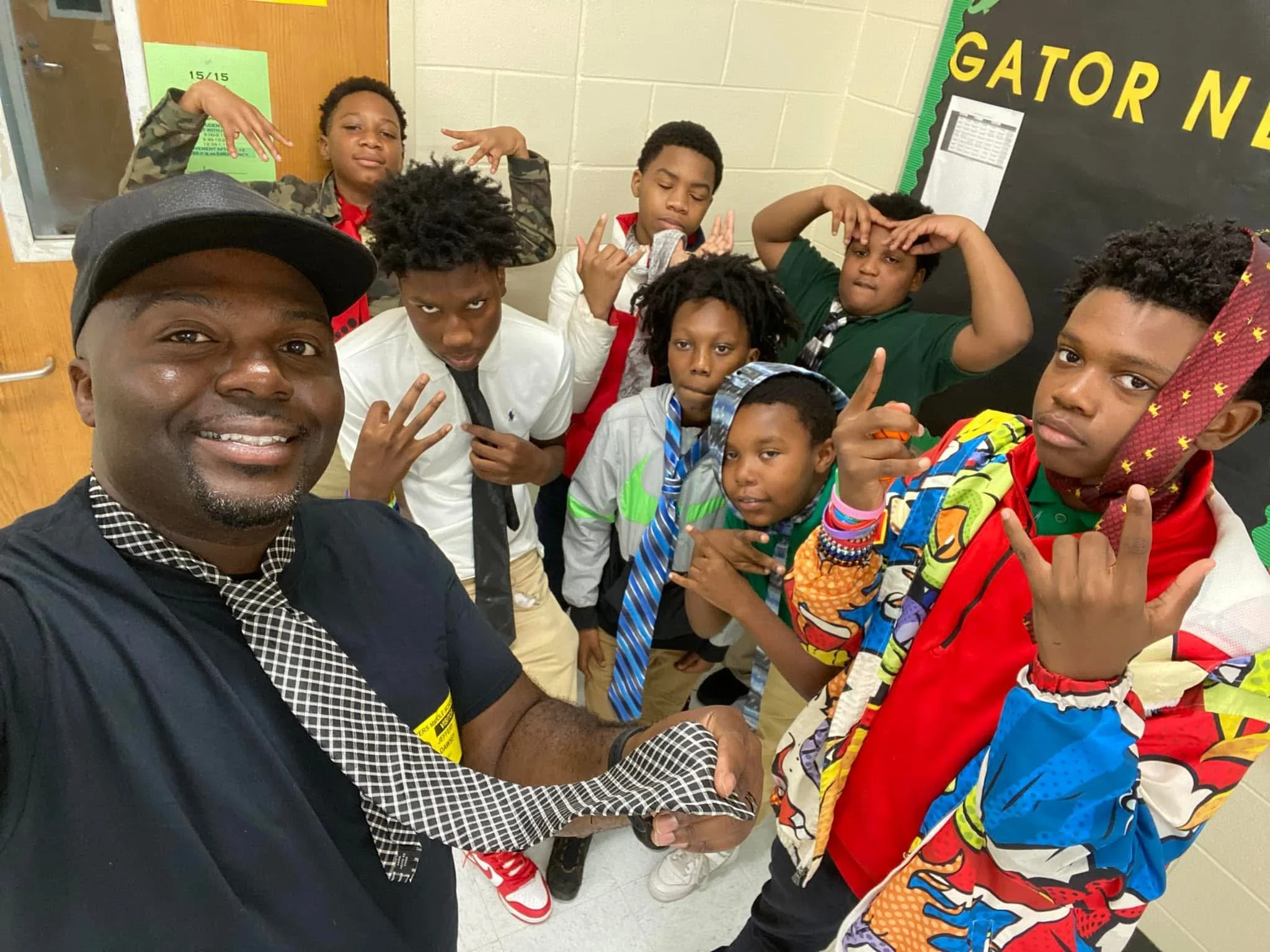 A group of school children and an adult taking a selfie in a classroom. The children are dressed in casual and school attire, making various hand gestures and facial expressions. The adult, wearing a black cap and shirt, is smiling at the camera.