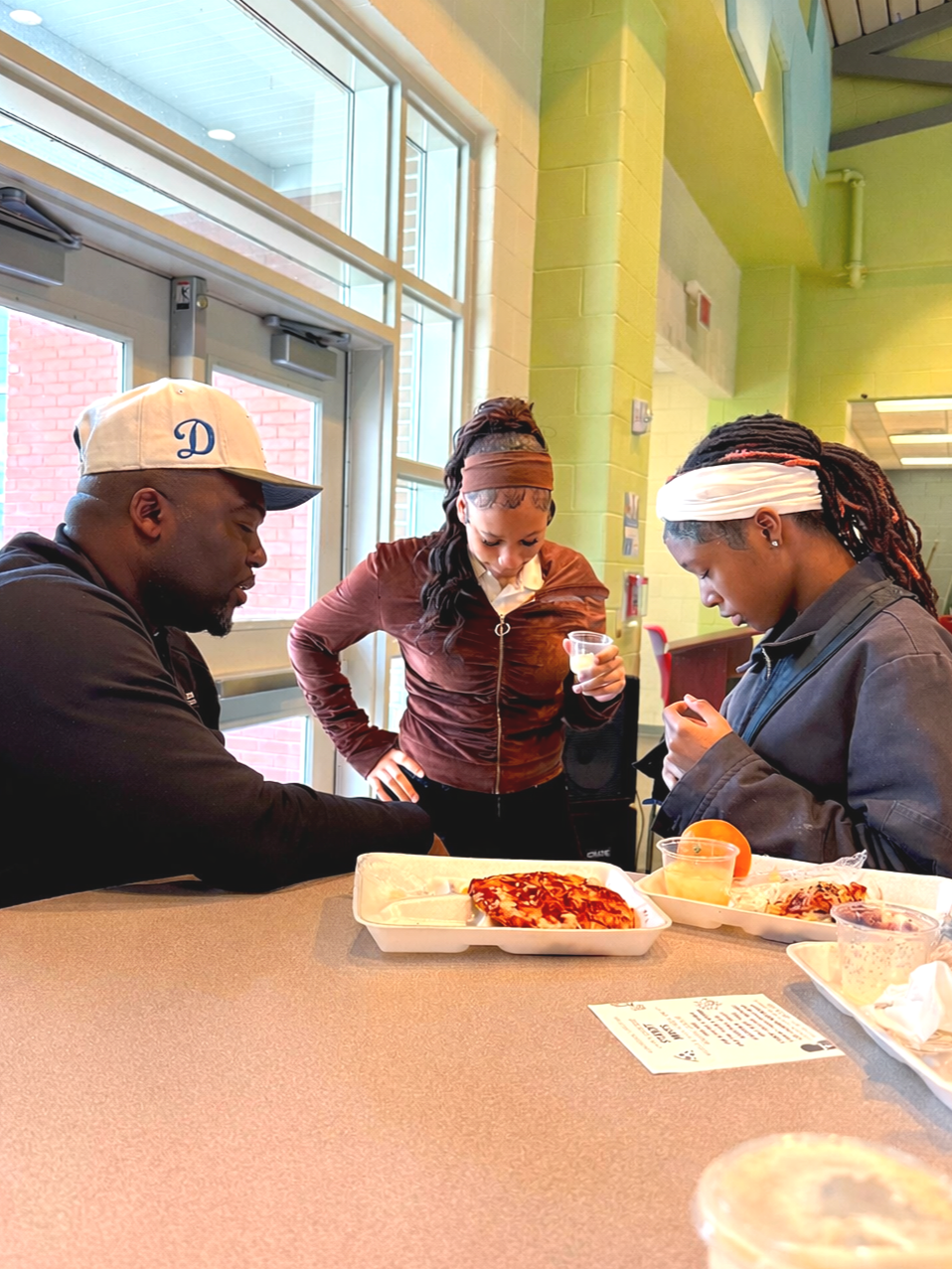 Three people standing and sitting around a table in a brightly lit indoor space, looking at food containers with pizza and snacks, with a window and green walls in the background.
