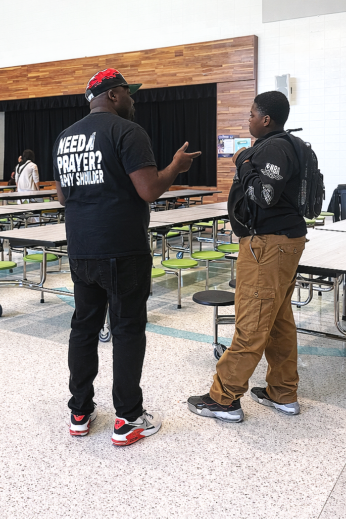 Two young men engaged in conversation in a cafeteria or dining hall, with tables and chairs around them and a black curtain behind.