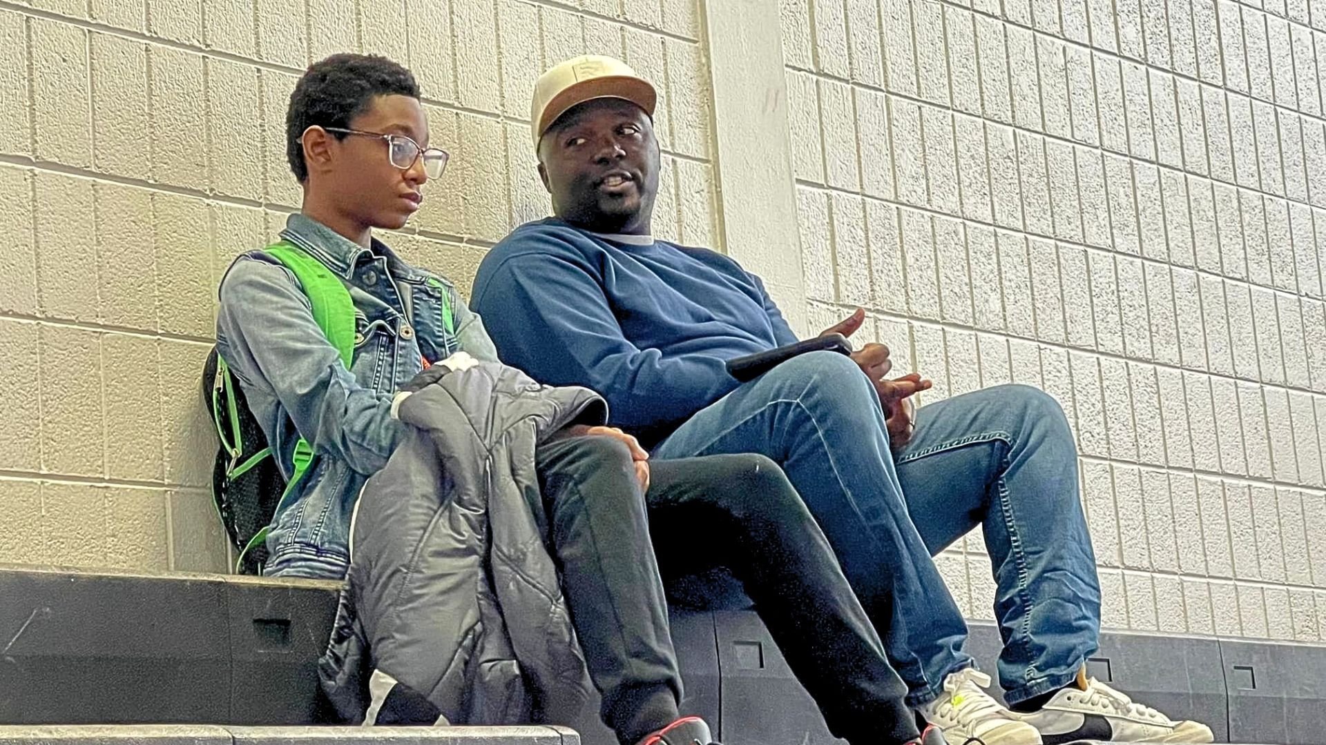 A young woman with short hair, glasses, and a denim jacket is sitting on a bench next to a man in a blue sweatshirt, jeans, and a tan cap, both are leaning against a brick wall in a public space.