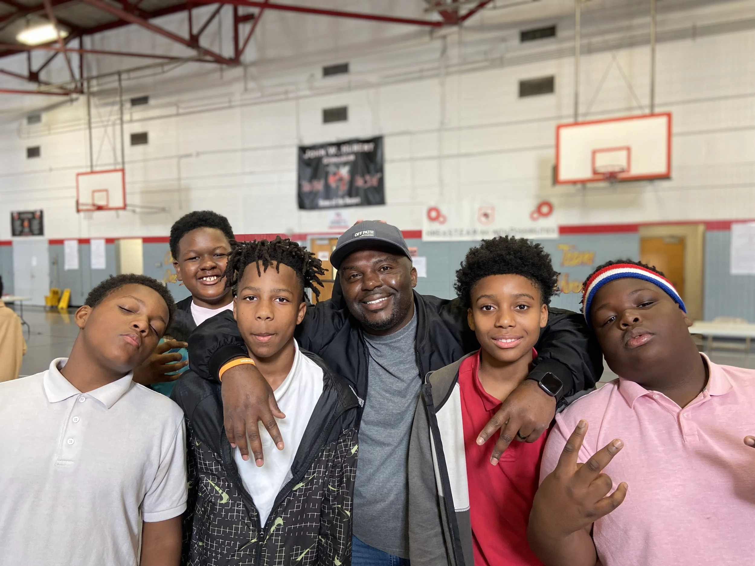 A group of six boys and one adult man posing together inside a gymnasium, smiling and making gestures. The background includes basketball hoops and gymnasium walls.
