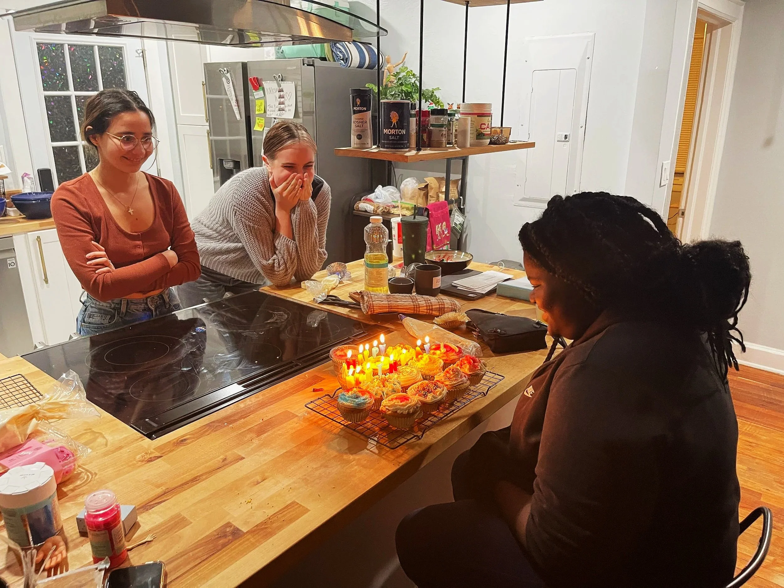 Three women gather around a kitchen island, with one woman sitting and the other two standing, as they celebrate a birthday with lit candles on cupcakes. The woman sitting has her head slightly bowed, and the two women standing smile joyfully.