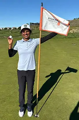 A woman standing on a golf course with a golf flag, holding a golf ball in her right hand and smiling at the camera.