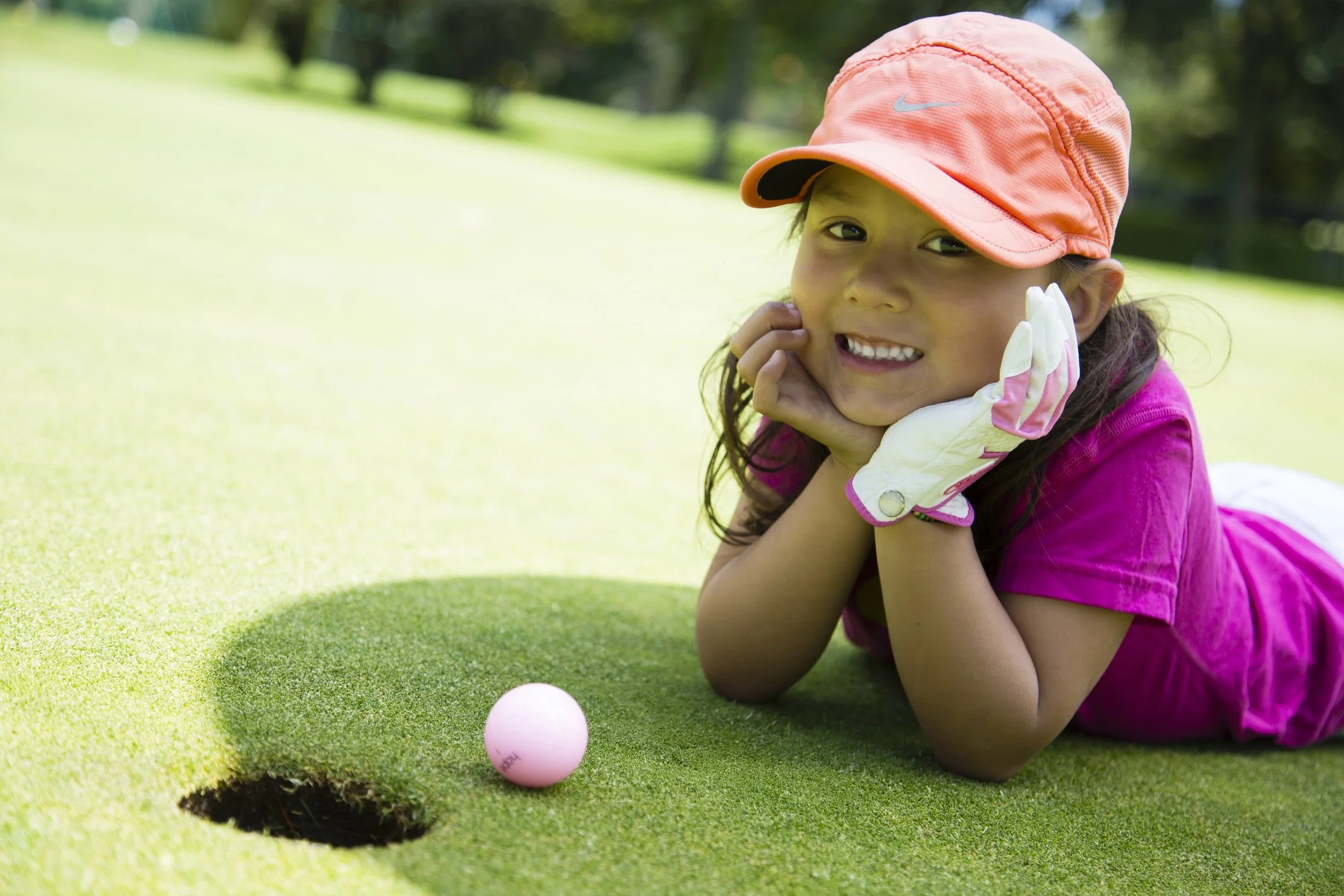 A young girl wearing a pink shirt, orange cap, and golf glove lying on a golf course, smiling, next to a pink golf ball near a hole.