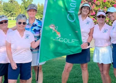 Group of five women in golf attire standing on a golf course with a green flag that says 'GOLF'. They are smiling with trees and flowers in the background.
