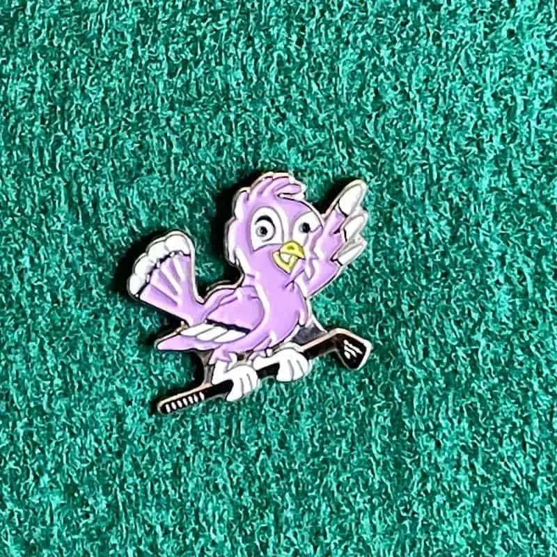 Cartoon-style purple bird with a yellow beak and eyes, perched on a black object, set against a textured green background.