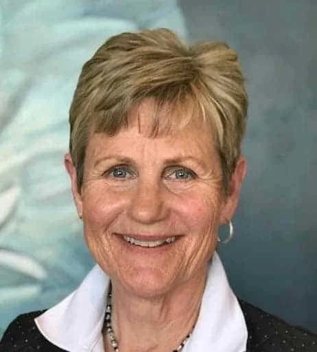 A smiling middle-aged woman with short, light brown hair, wearing a white collared shirt, black blazer, and pearl necklace.