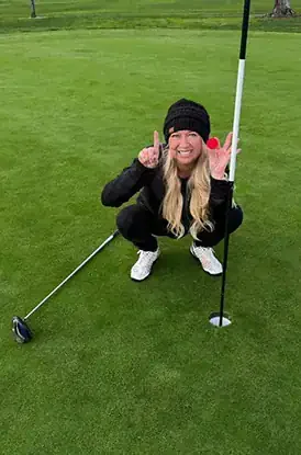 A woman crouching on a golf green, holding a golf flagstick with her left hand, smiling and making a peace sign with her right hand, dressed in black with a black beanie.