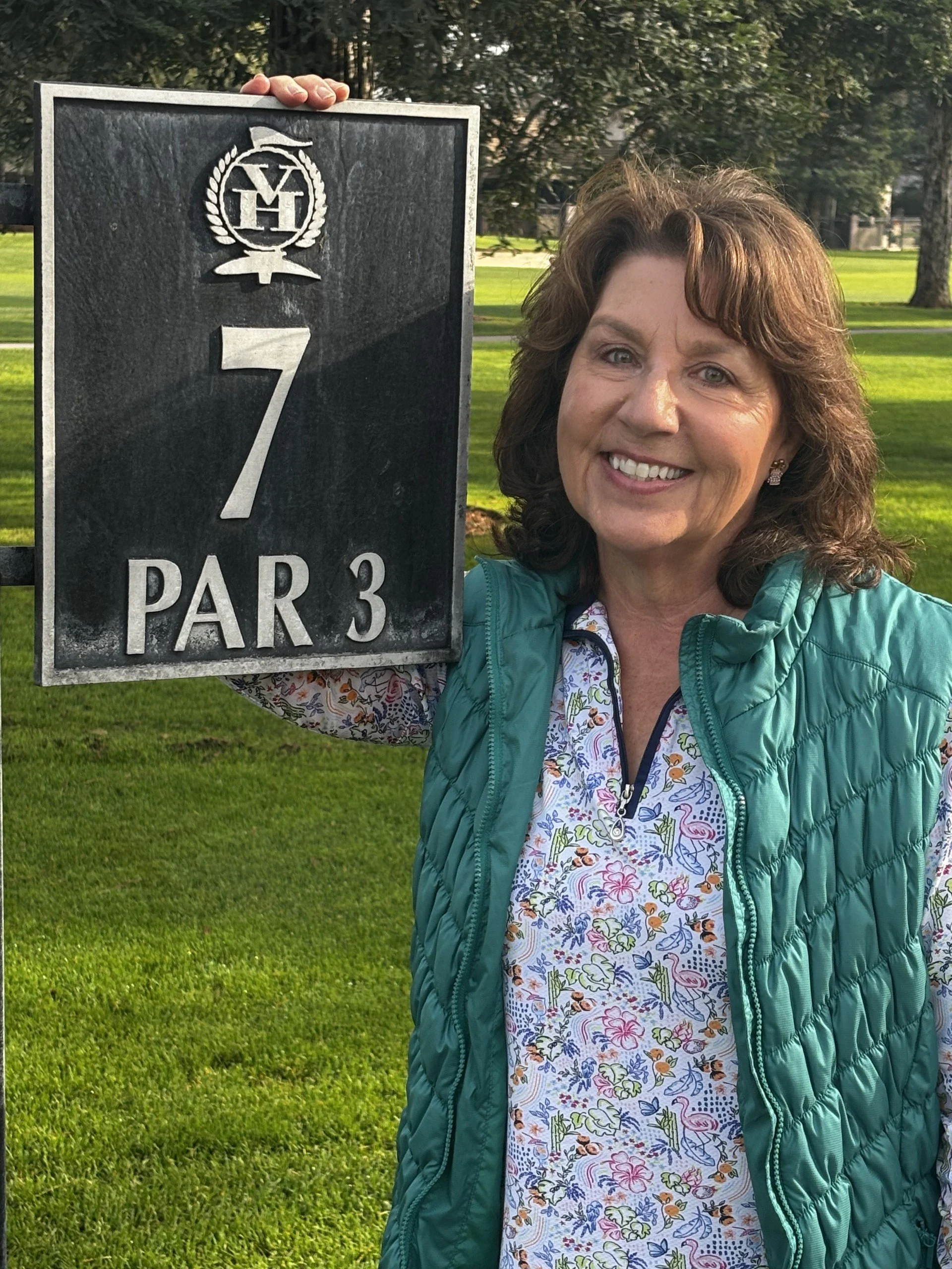 A woman smiling and holding a golf sign on a golf course with trees and grass in the background.