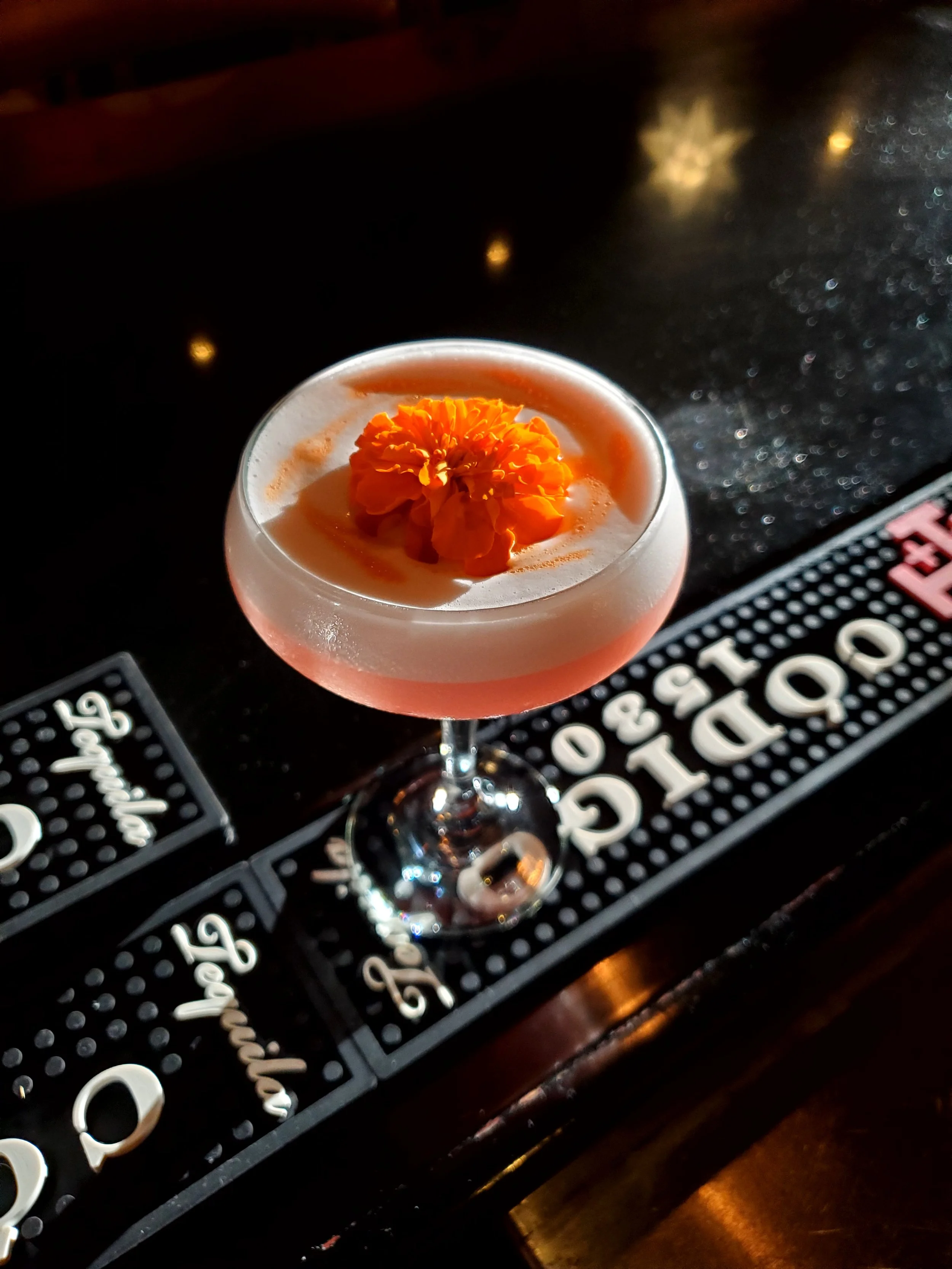A cocktail glass with an orange flower garnish on top of a creamy pink drink, placed on a bar counter.