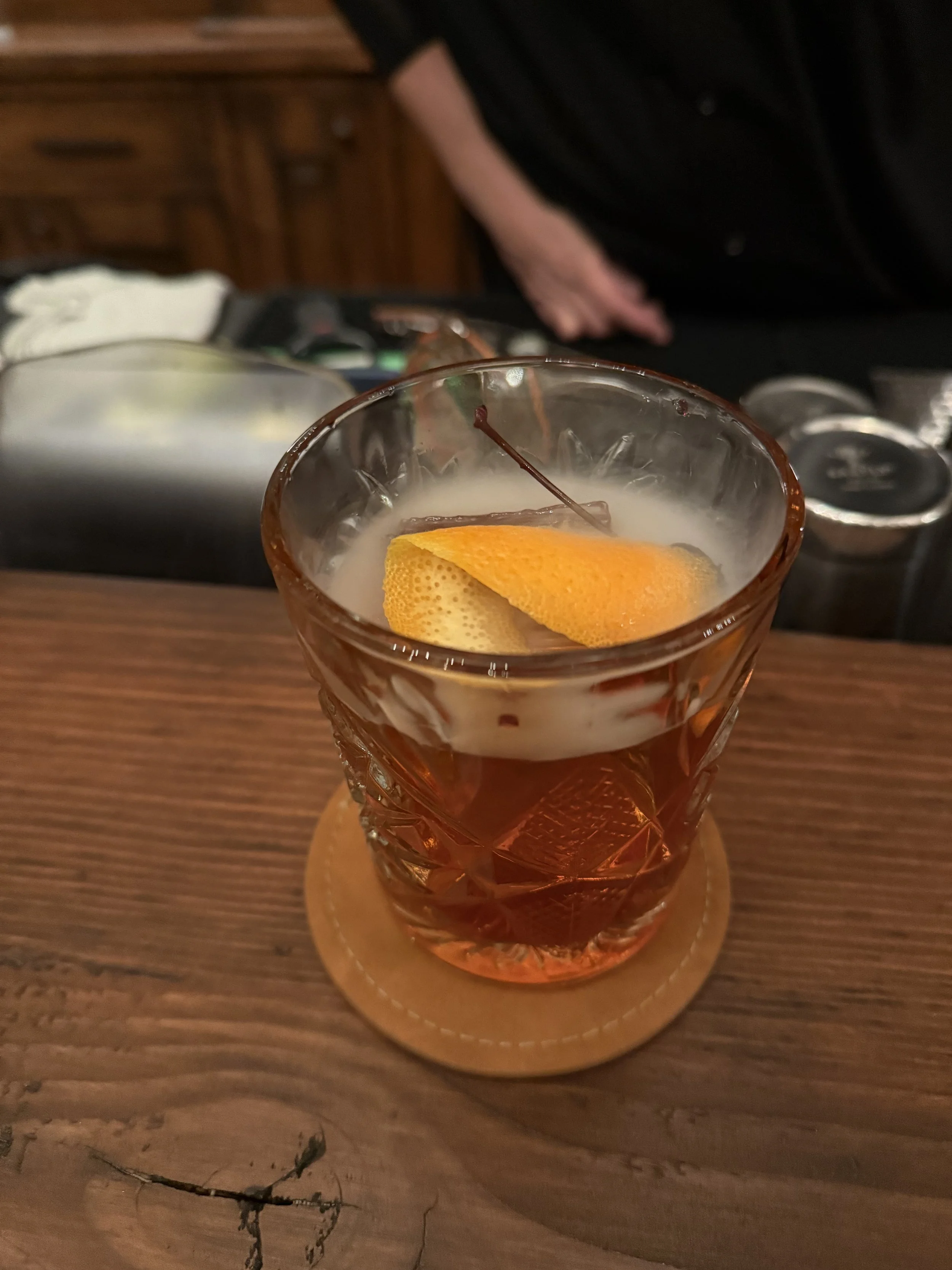 A glass of cocktail with a lemon peel garnish on a wooden table, with a person in dark clothing in the background.
