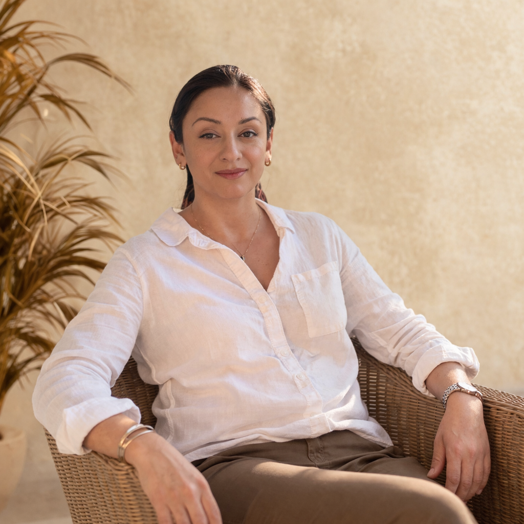 A woman sitting in a wicker chair with a relaxed posture, wearing a white shirt and brown pants, indoors with a beige wall and a potted plant in the background.