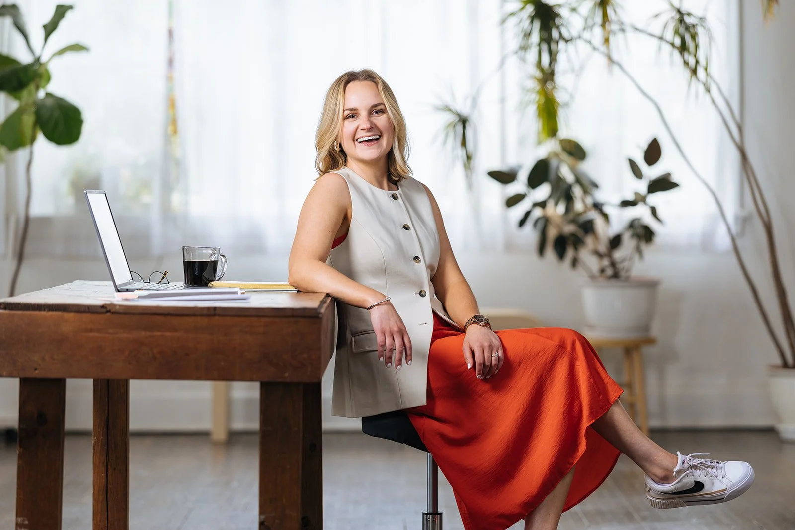 A smiling woman with blonde hair sitting on a chair in a bright room with plants in the background. She is wearing a white sleeveless vest, a red skirt, and white sneakers. There is a wooden table with a laptop, a pair of glasses, a cup of coffee, and notebooks on it.