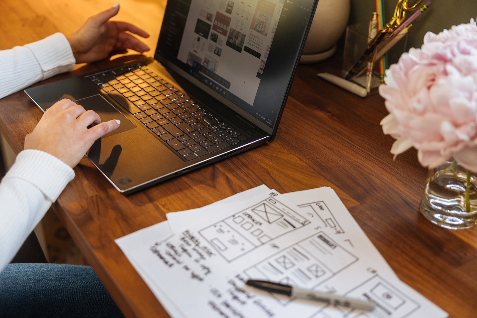 Person working on a laptop at a wooden desk with design sketches, flower vase, and office supplies.