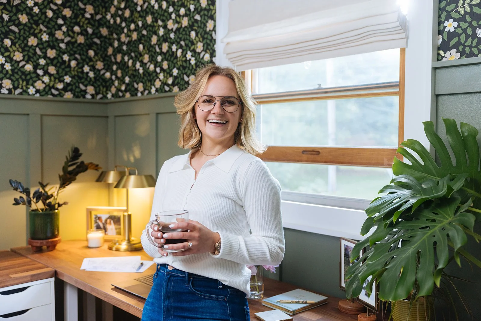 A woman with blonde hair, glasses, and a white sweater smiling while holding a cup of coffee in a cozy, decorated room with green walls, a wooden desk, potted plants, and framed photos.