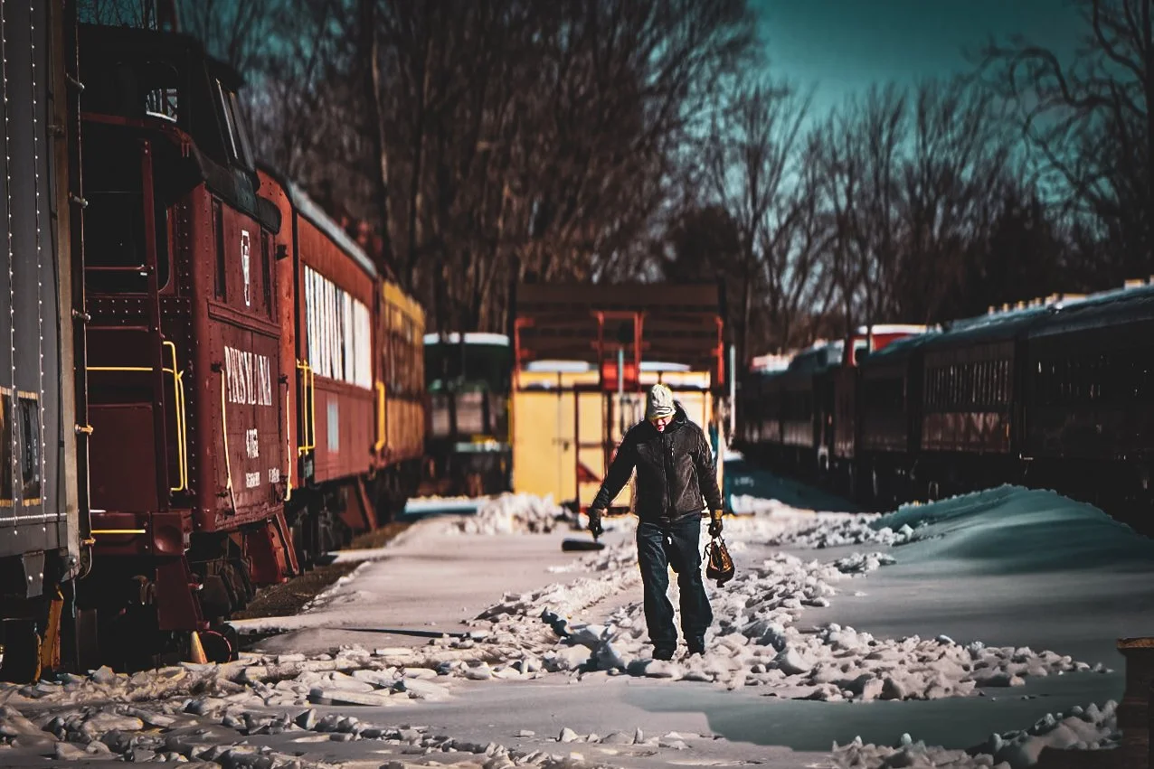 Train Yard Documentary Scene
Documentary photograph of a person walking through a snowy rail yard beside historic train cars.