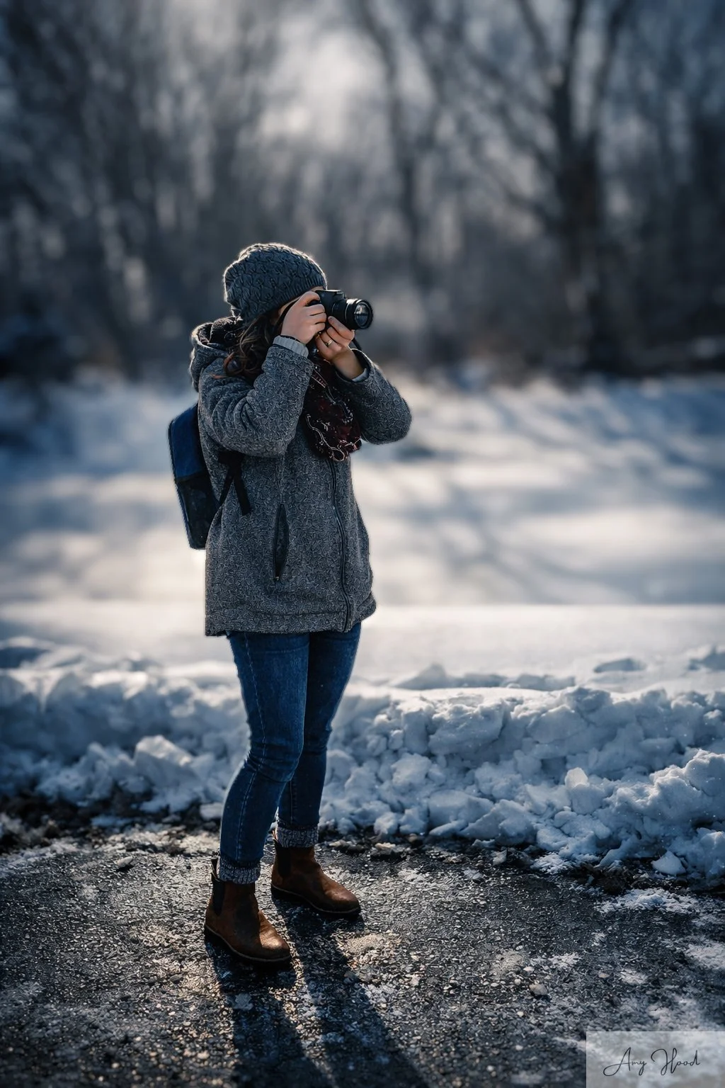 A woman standing outdoors in a snowy landscape, taking a photograph with a camera. She is dressed in a gray coat, knitted beanie, jeans, and brown boots, with a backpack on her back. The background features snow-covered ground and leafless trees.