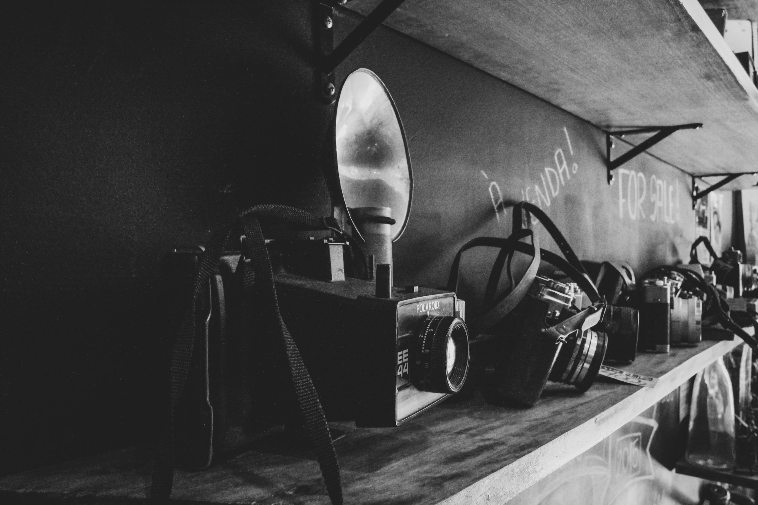 Black and white photo of a wooden shelf displaying vintage cameras of various styles and sizes, some with straps, with handwritten text on the wall behind reading 'BRAND! FOR SALE!'