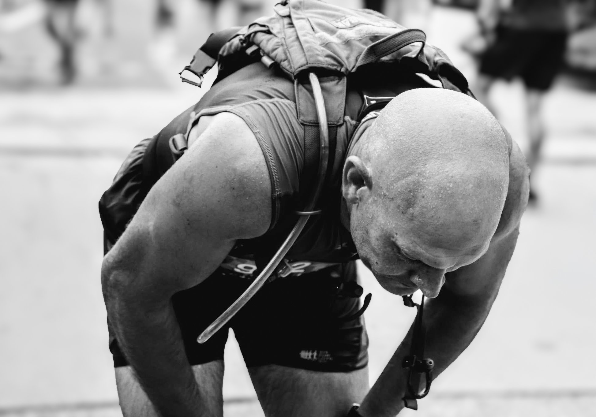A male athlete with a shaved head hunched over, exhausted, with sweat on his face and arms, wearing sports gear and a hydration pack, likely after a race.
