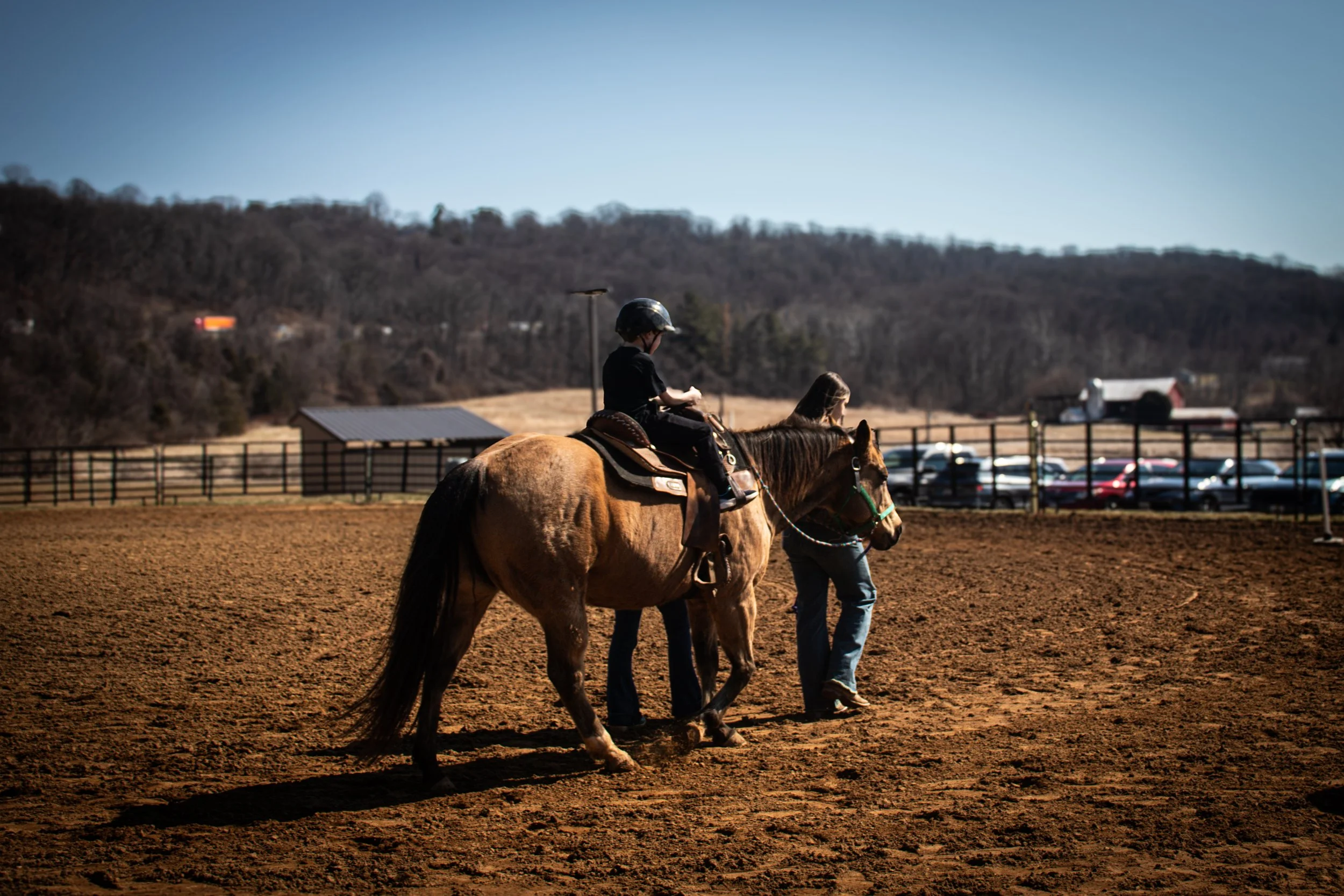 a young boy has his first pony ride as the trainer is leading the horse across a sunlit field, capturing a quiet moment of rural life.