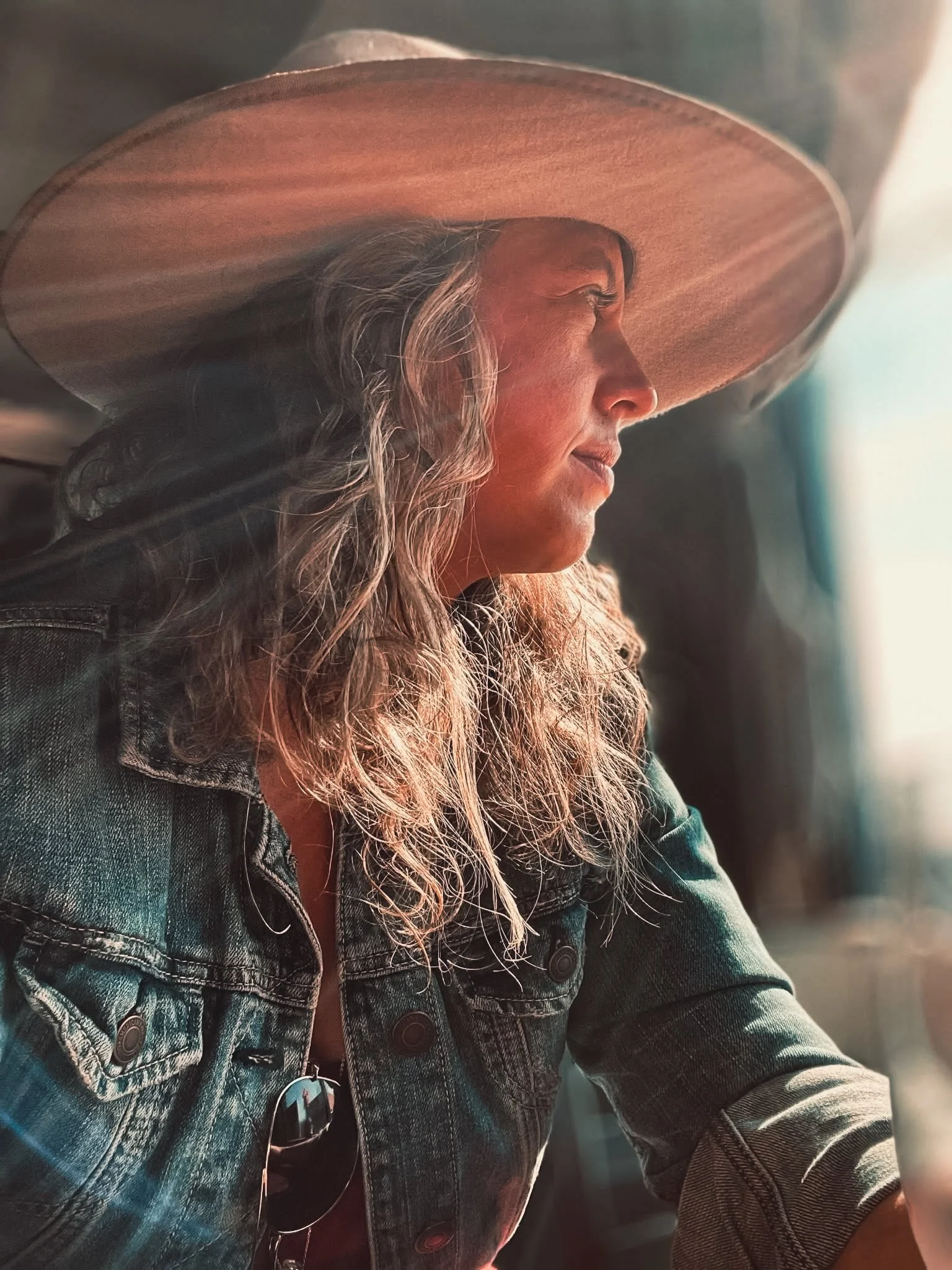Side profile of a woman with long wavy hair wearing a wide-brimmed hat and sunglasses, sitting with a denim jacket, looking towards the light.