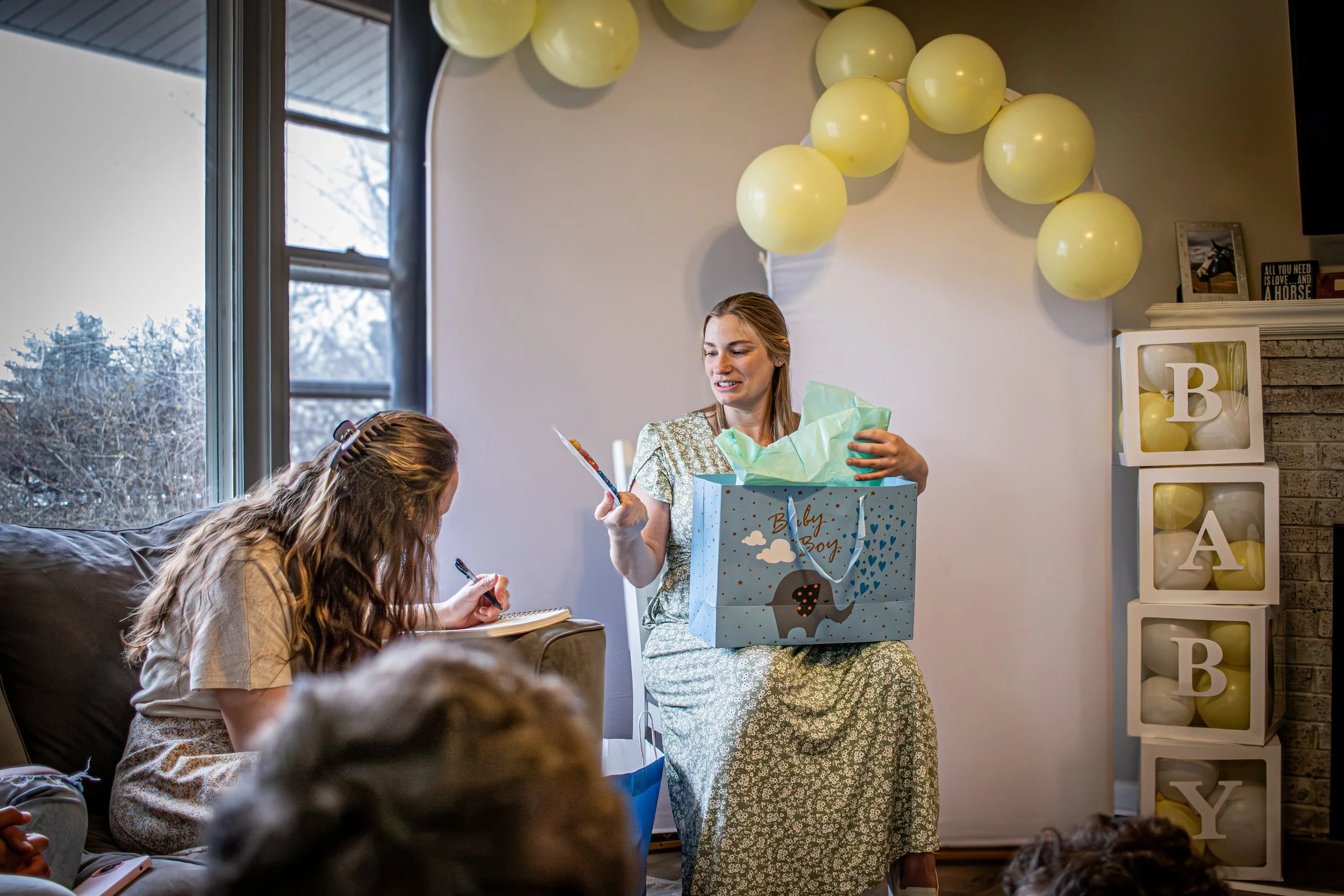 Mother-to-be opening a baby shower gift while seated in front of yellow balloon decorations as guests watch during a cozy indoor celebration
