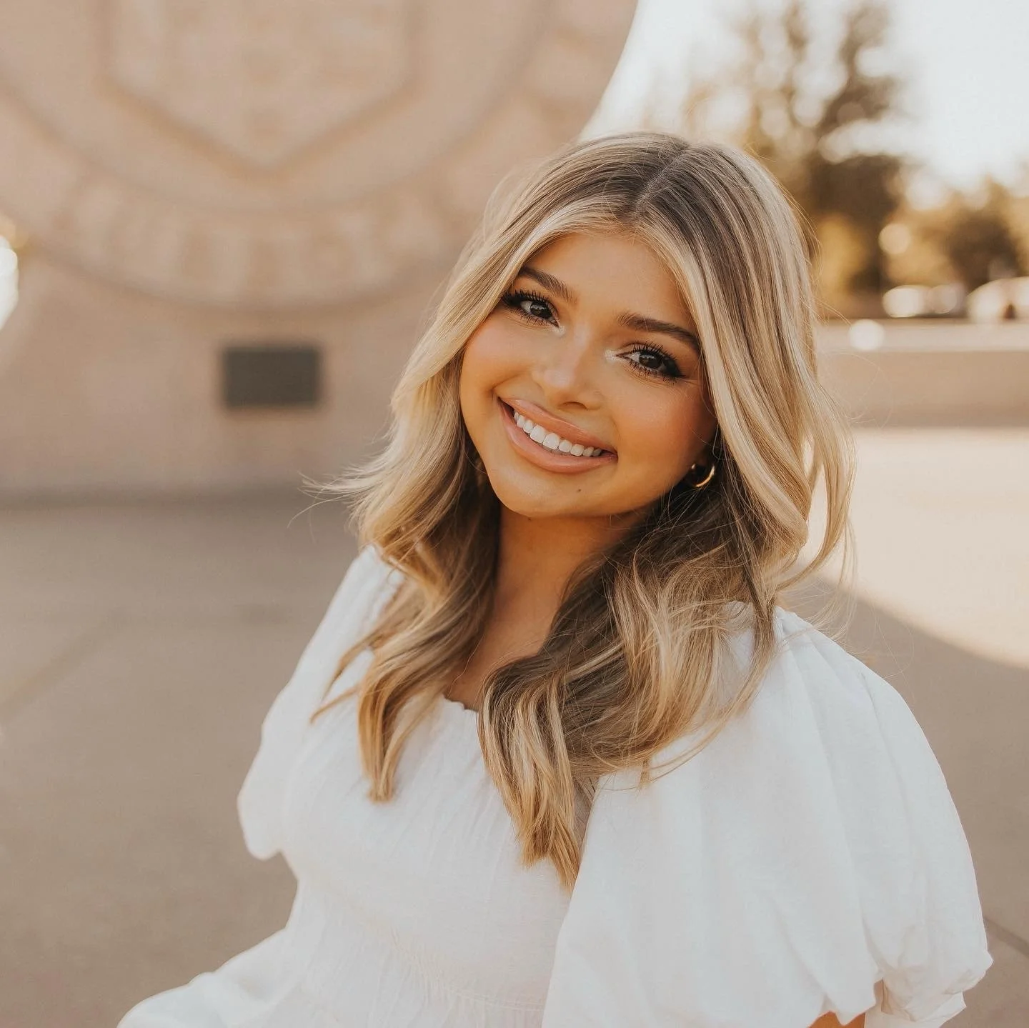 Smiling woman with blonde hair in loose waves, wearing white blouse, outdoors during golden hour.