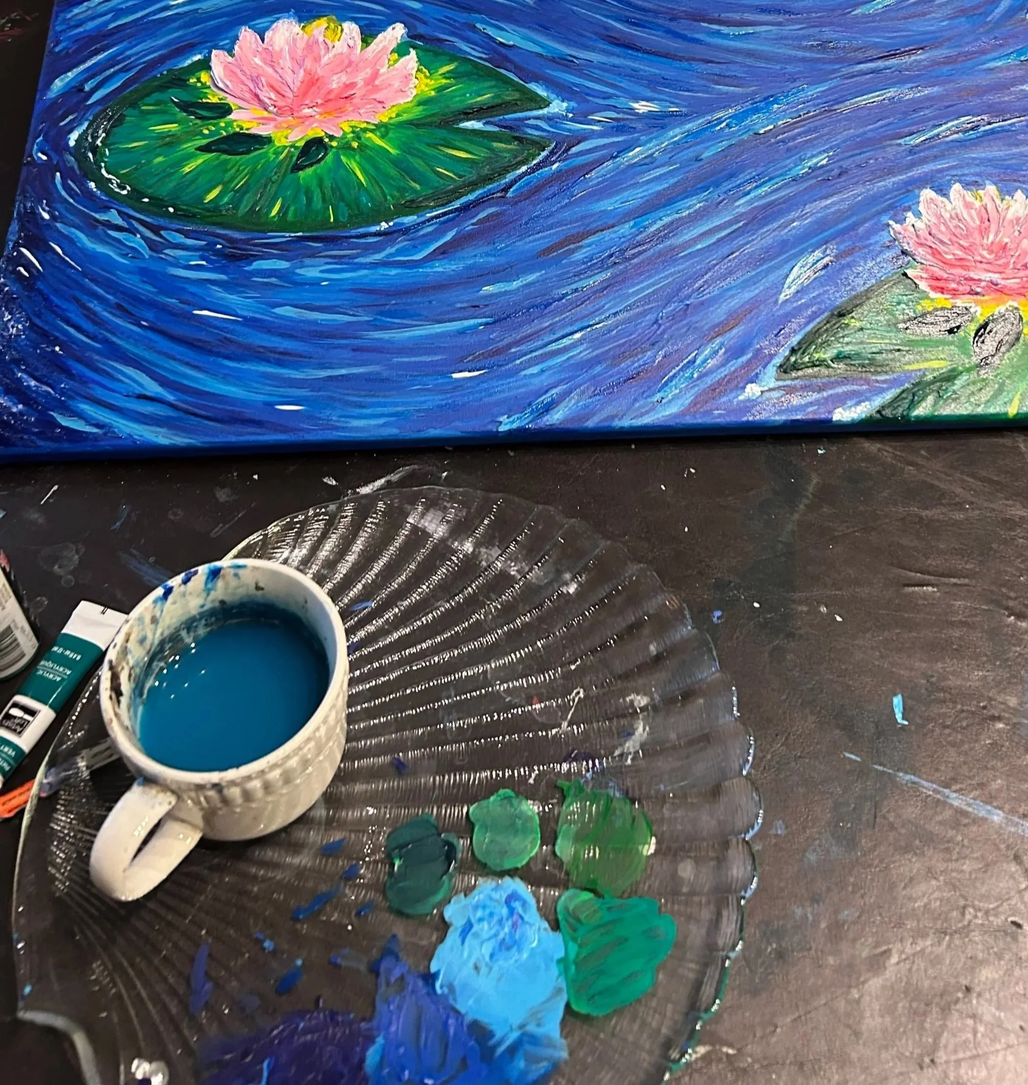 A painting of two pink water lilies with green lily pads on a blue water background, with artists' paint and tools in the foreground.