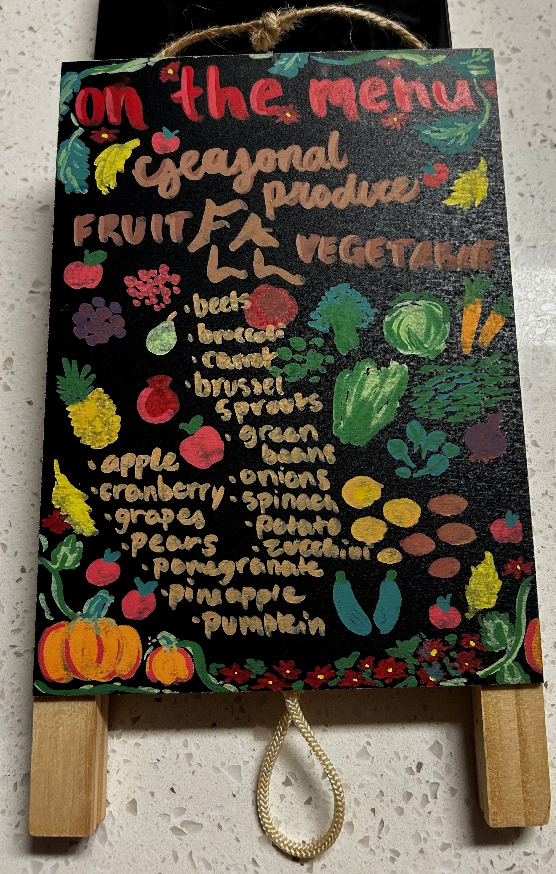 A decorative black chalkboard with colorful drawings of vegetables, fruits, and flowers, listing produce items available on the menu, with a rope handle at the top and attached wooden pieces at the sides.