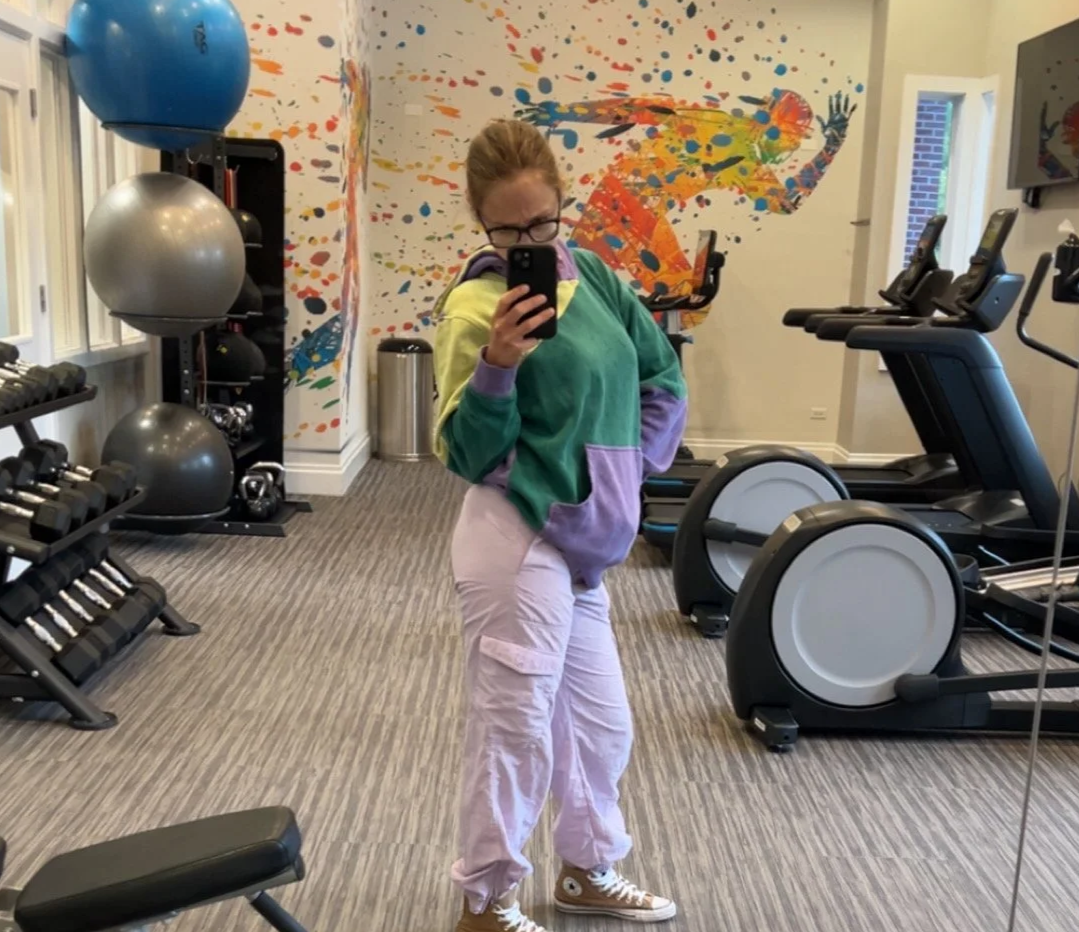 A woman taking a mirror selfie in a gym with colorful abstract elephant artwork on the wall, exercise equipment, and large exercise balls.
