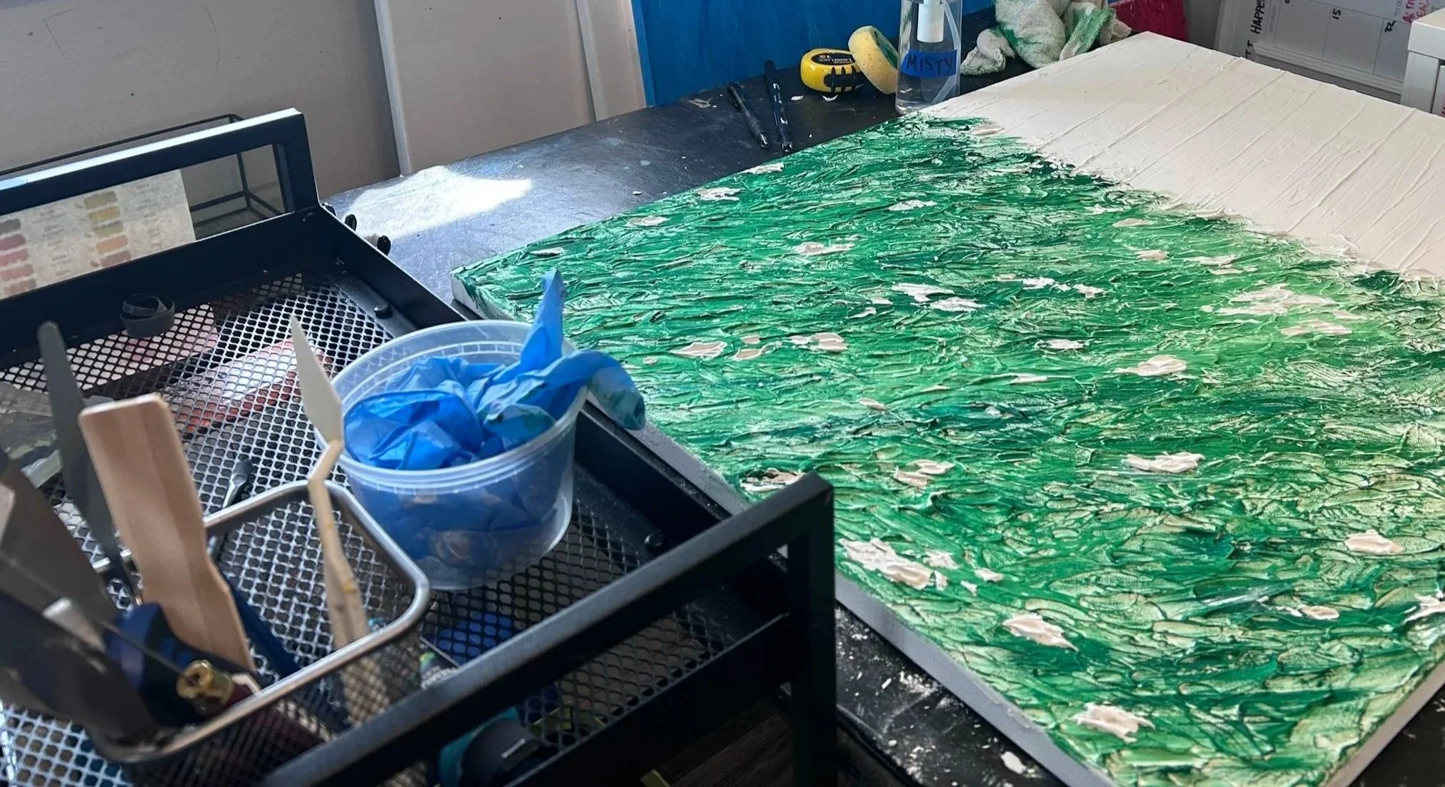 A workspace with a partially painted canvas in green and white, with art supplies including paint, brushes, and gloves on a table, in an art studio.