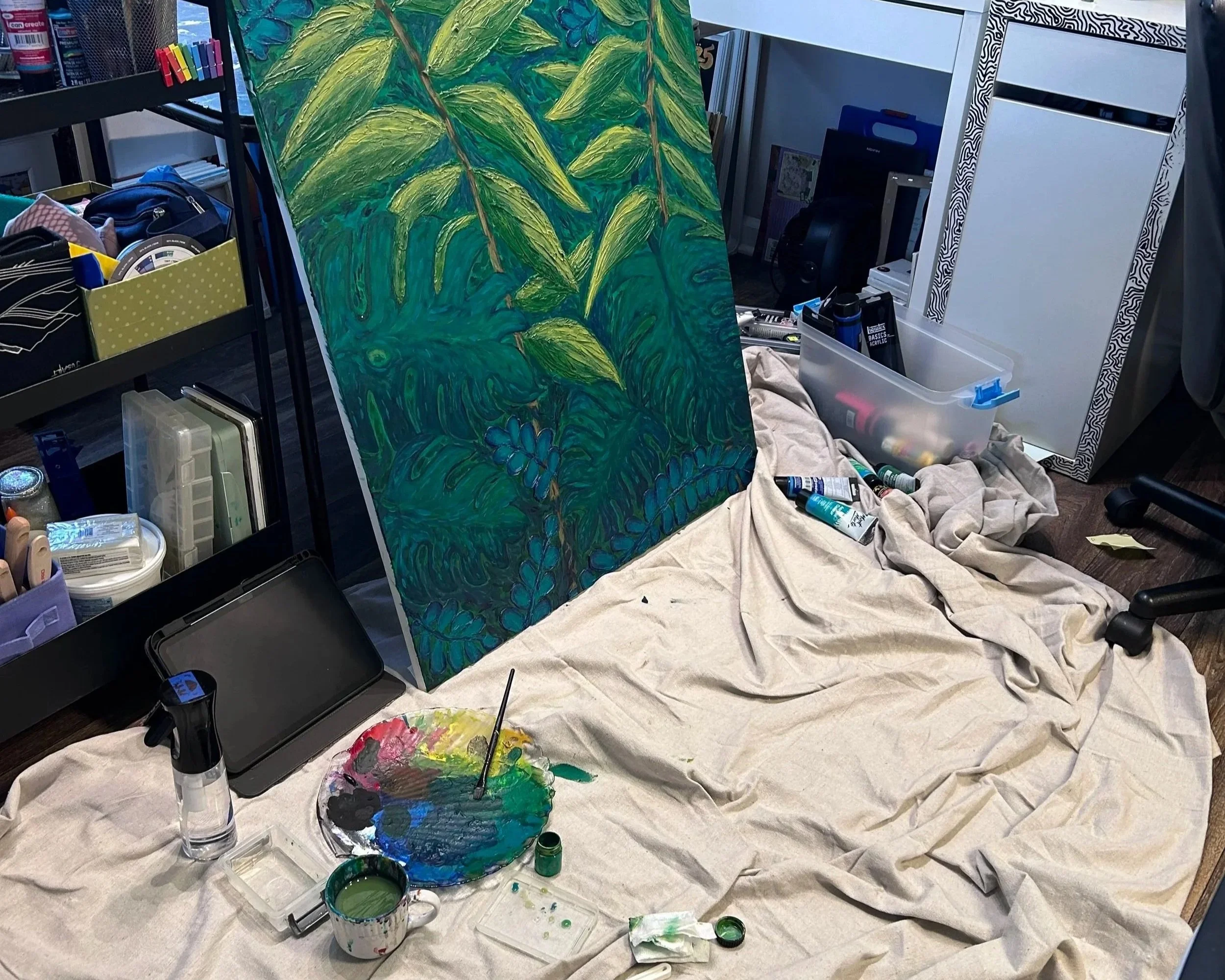 An art studio setup with a large canvas painted with green leafy foliage, acrylic paint supplies including open paint jars with green and other colors, brushes, a palette, and a in-progress painting. Art supplies, mirrors, and shelves with various items are visible in the background.