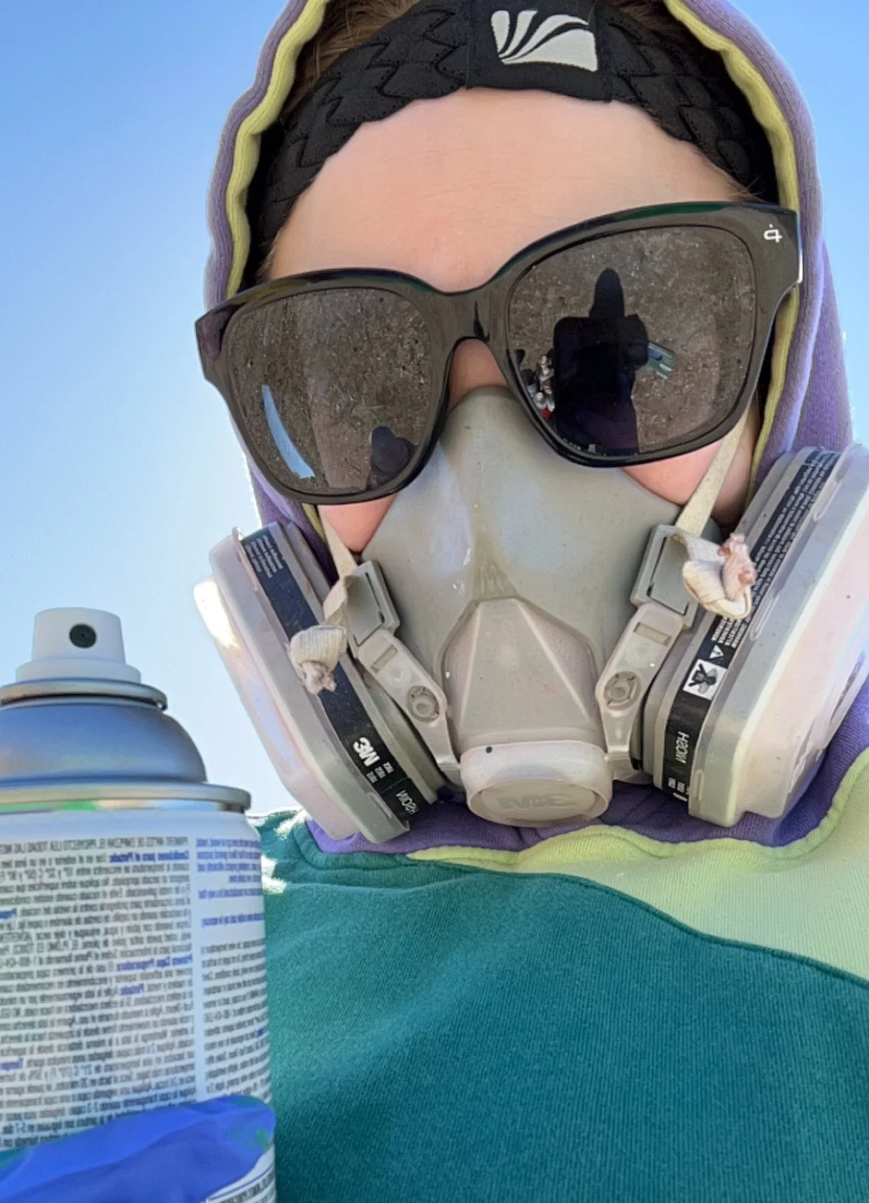 Close-up of a person wearing sunglasses, a black headband, a dust mask, and a purple hoodie, with a blue sky in the background. Partially visible spray paint can and a can of aerosol spray are in the foreground.