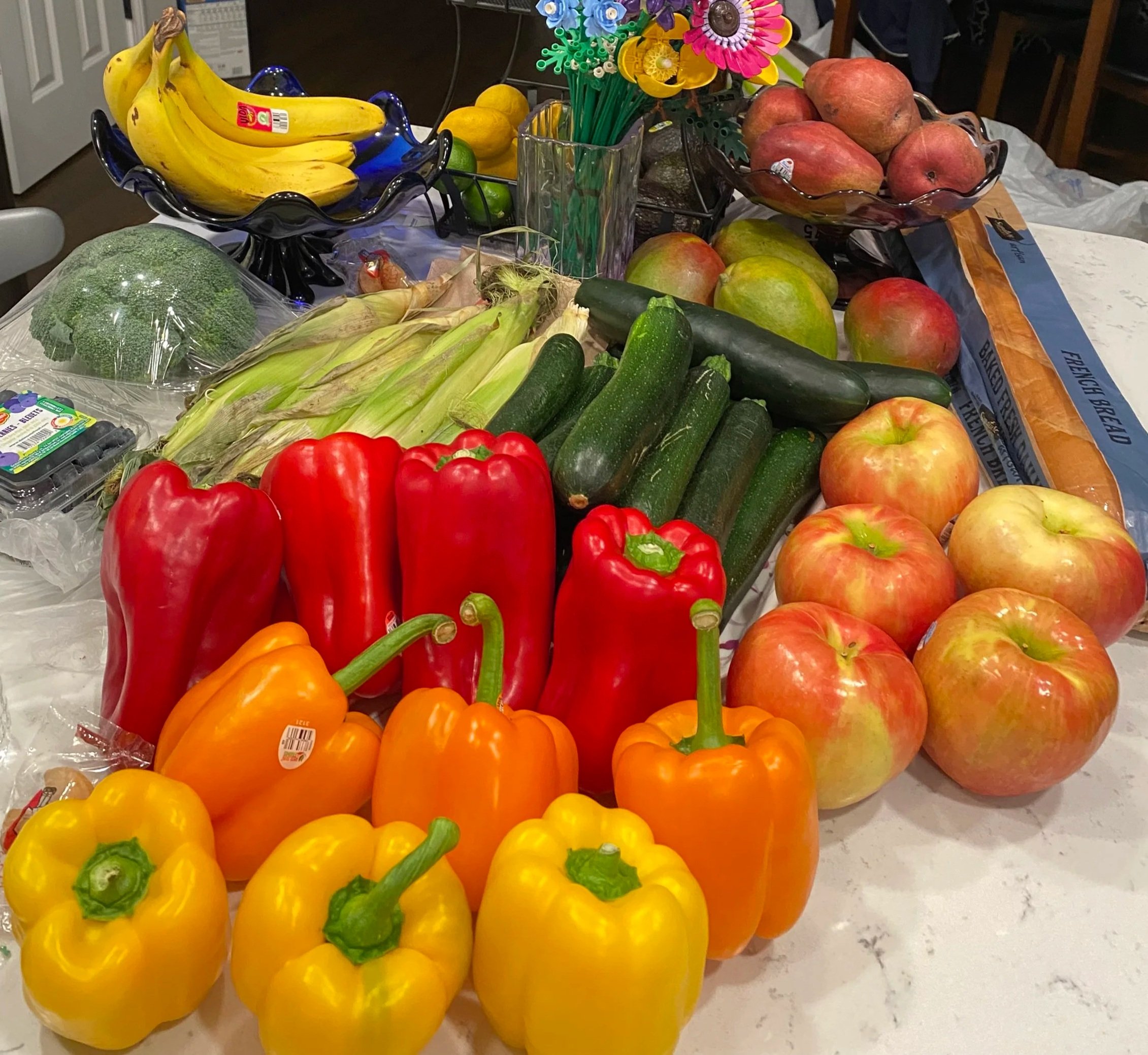 A table filled with fresh produce including bananas, bell peppers, tomatoes, cucumbers, apples, and broccoli, with a vase of colorful artificial flowers in the background.