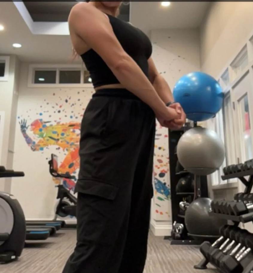 A person standing in a gym, holding their hands together, with gym equipment including exercise balls and dumbbells in the background.