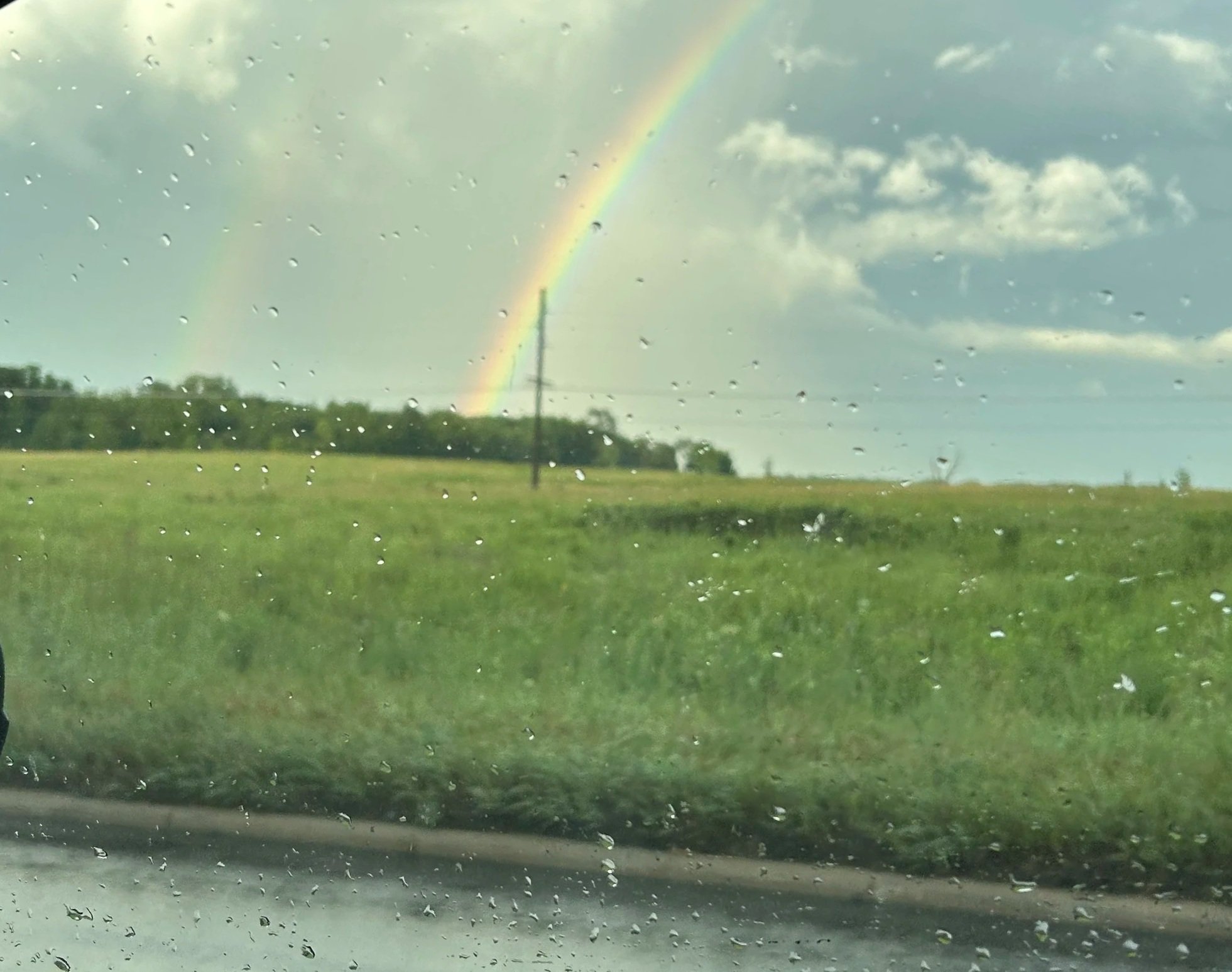 Rain pouring on a window with raindrops, a green field, trees, a utility pole, and a rainbow in the cloudy sky.