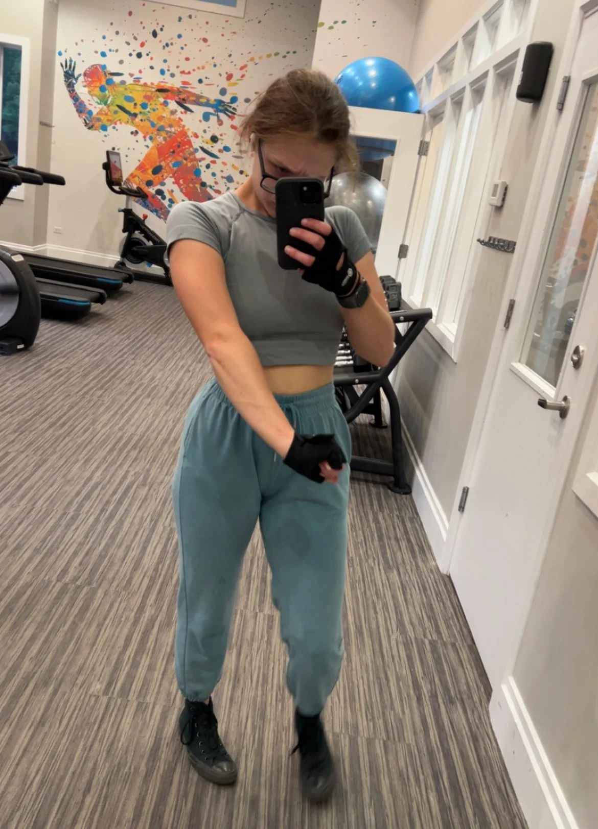 A woman in workout attire taking a mirror selfie in a gym, wearing a gray crop top, teal joggers, and black sneakers, with colorful wall art, exercise equipment, and large exercise balls in the background.