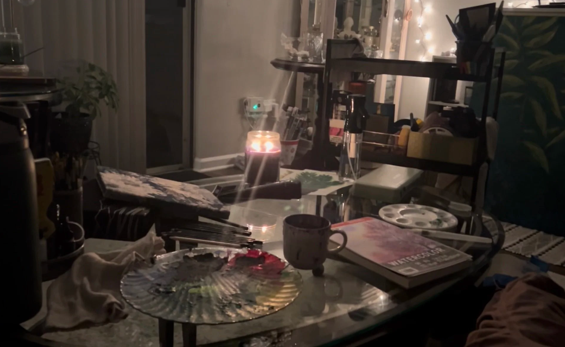 A cluttered table with a lit candle, a cup, a paint palette, magazines, and art supplies, with a background of shelves filled with various items and string lights.