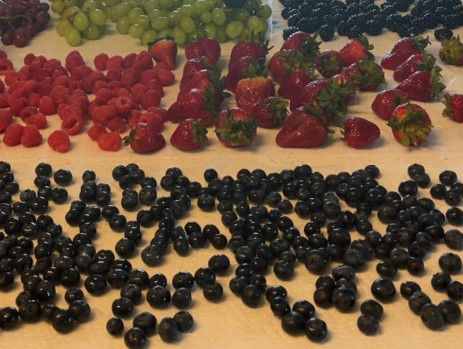 Assorted fresh berries on a wooden surface, including green grapes, blackberries, strawberries, raspberries, and blueberries.