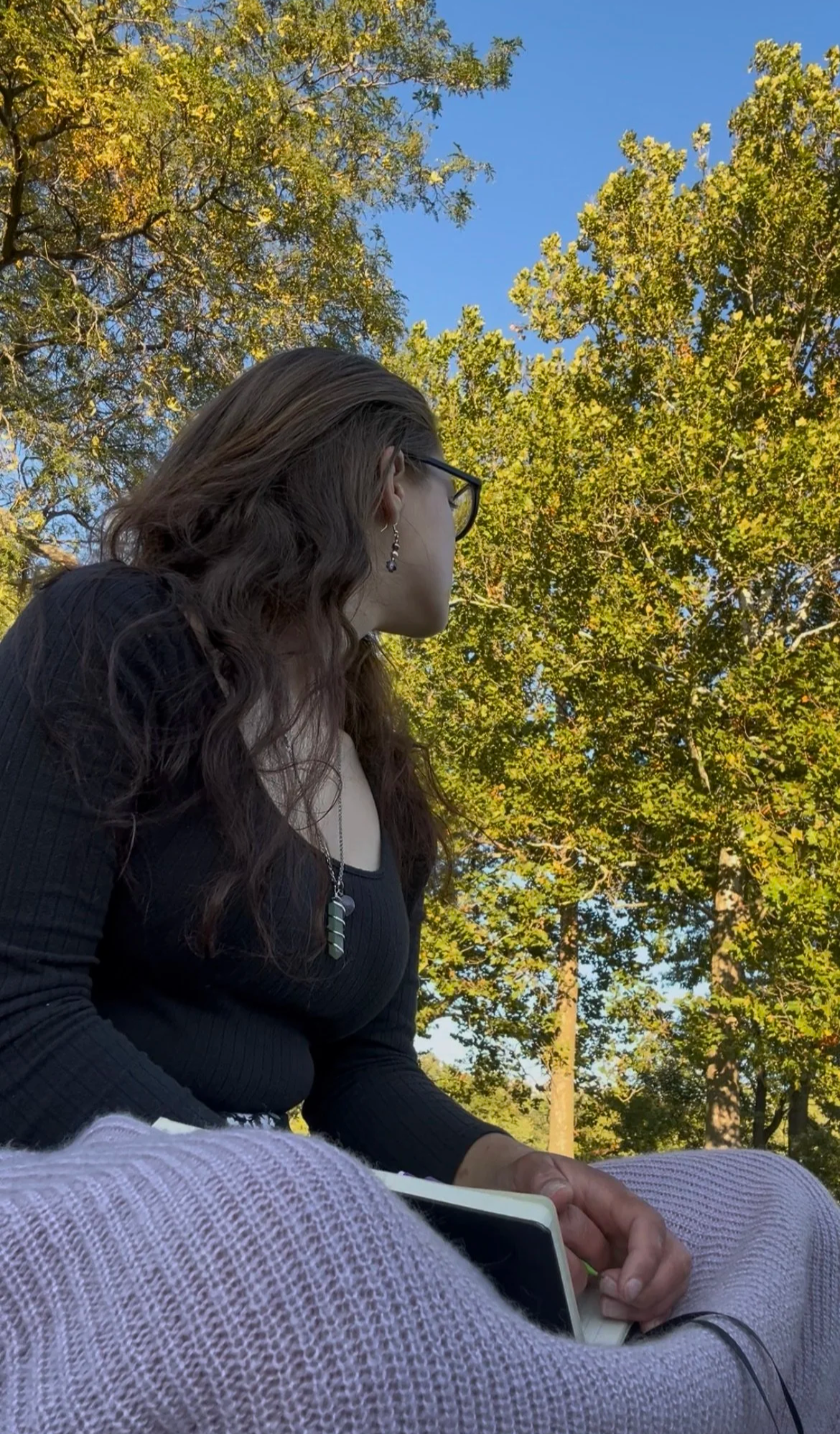 A woman with wavy brown hair, wearing glasses, earrings, and a black shirt, sits outdoors surrounded by green trees and a clear blue sky, holding a book or notebook.