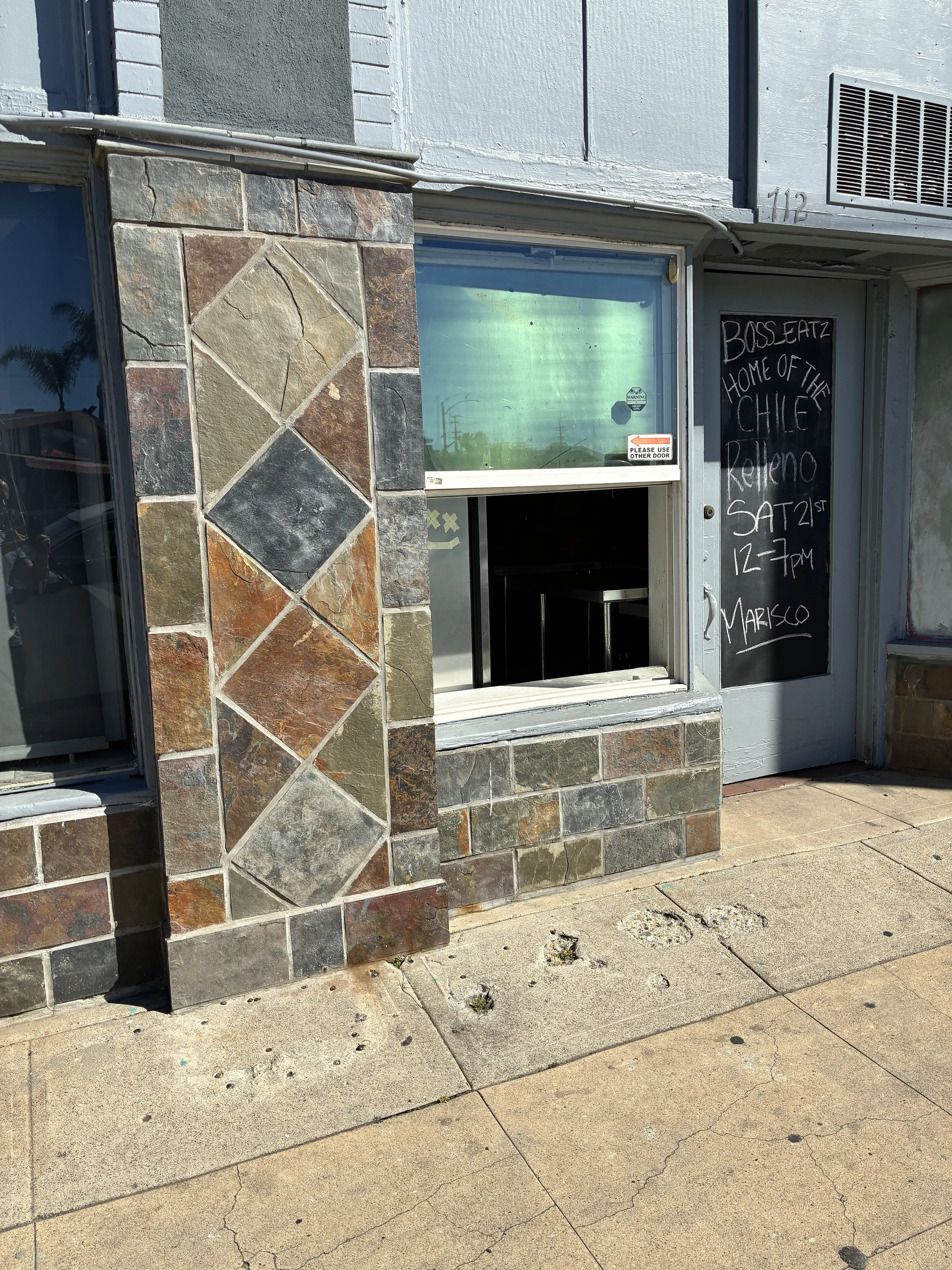 A restaurant window with an open service hatch, next to a blackboard sign with handwritten event details, decorated with a stone wall and a sidewalk outside.