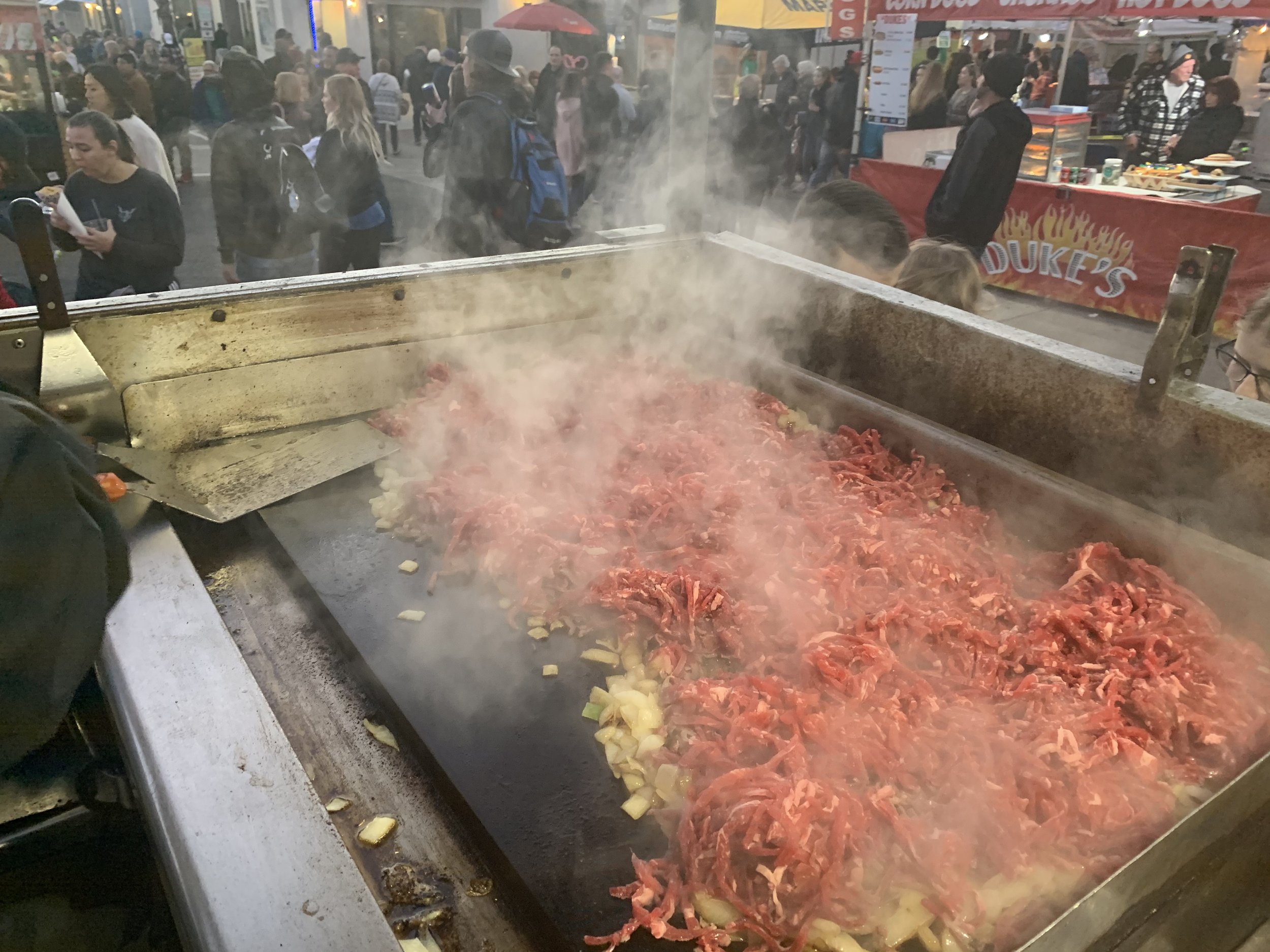 Large outdoor food stand cooking shredded beef with onions on a flat-top grill at a busy festival or fair. There are many people in the background, some waiting in line and others walking around.