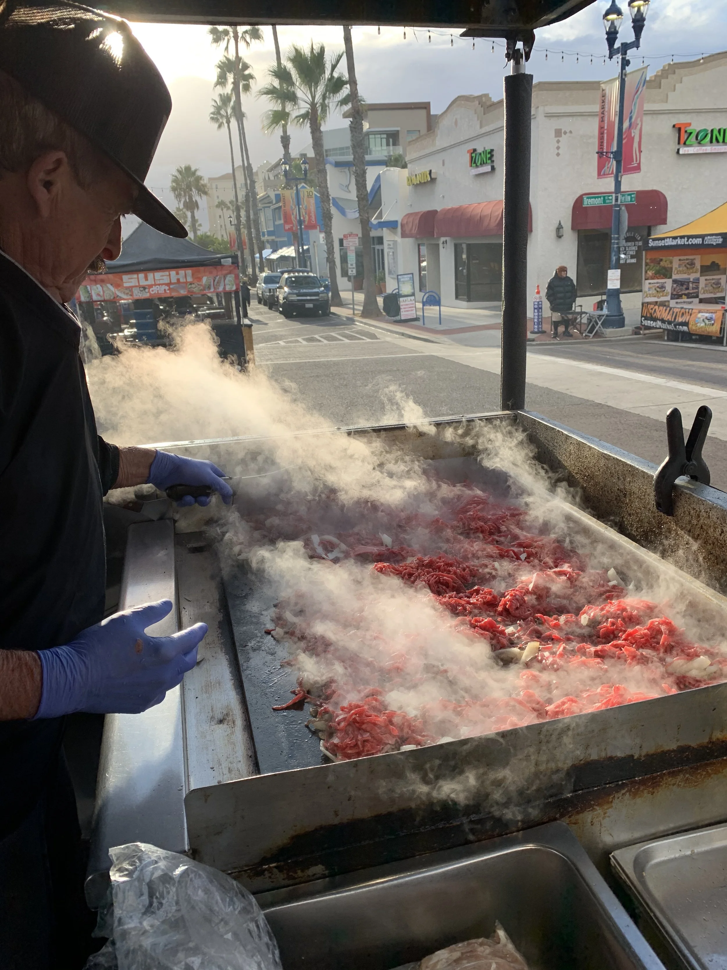A man wearing a cap and blue gloves cooking at a street food cart, grilling meat on a hot griddle with steam rising, with storefronts and palm trees in the background.