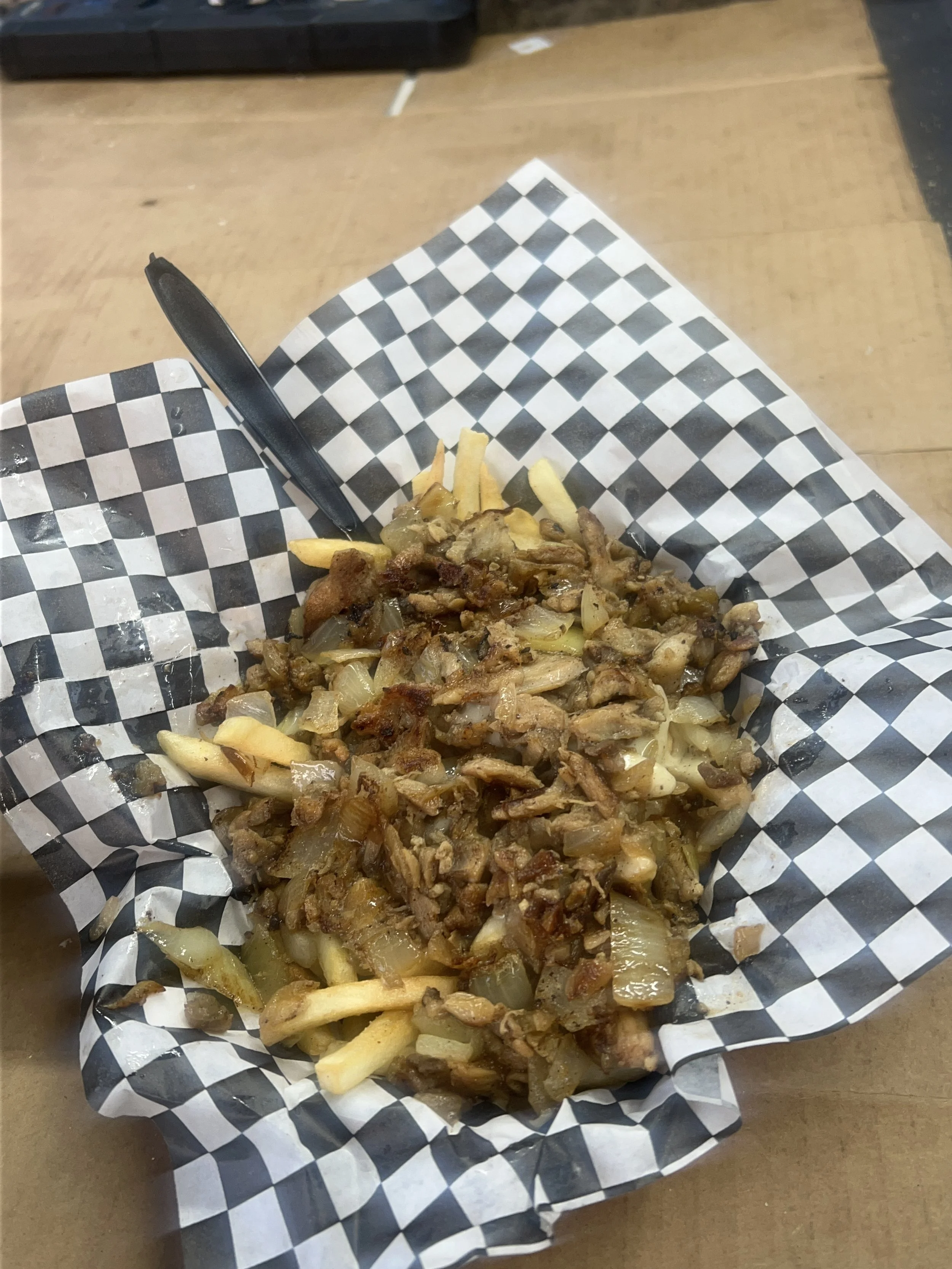 French fries topped with chopped grilled chicken, onions, and a sauce, served in a black and white checkered paper basket with a black plastic fork.